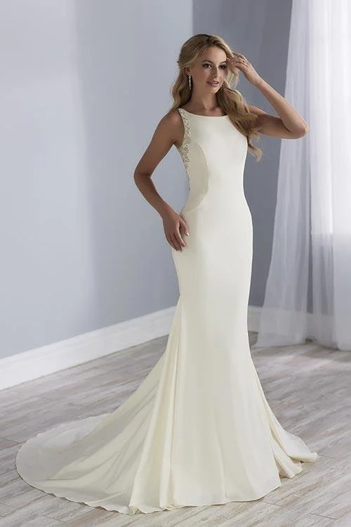 A woman in a fitted white wedding gown with lace details on the shoulders, standing indoors near a window with white curtains.