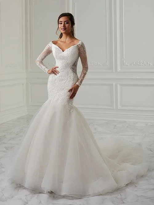 A woman in a white wedding dress with long lace sleeves, standing in a bright, elegant room with white walls and marble flooring.