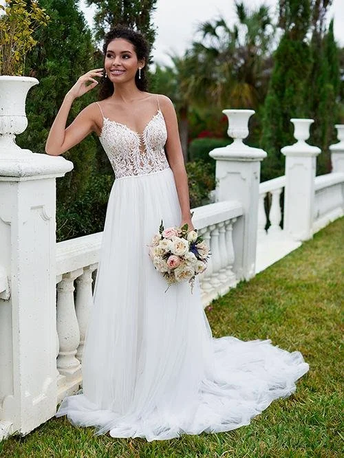 Young woman in a white wedding dress holding a bouquet of flowers, standing outdoors by a white decorative fence with lush green trees in the background.