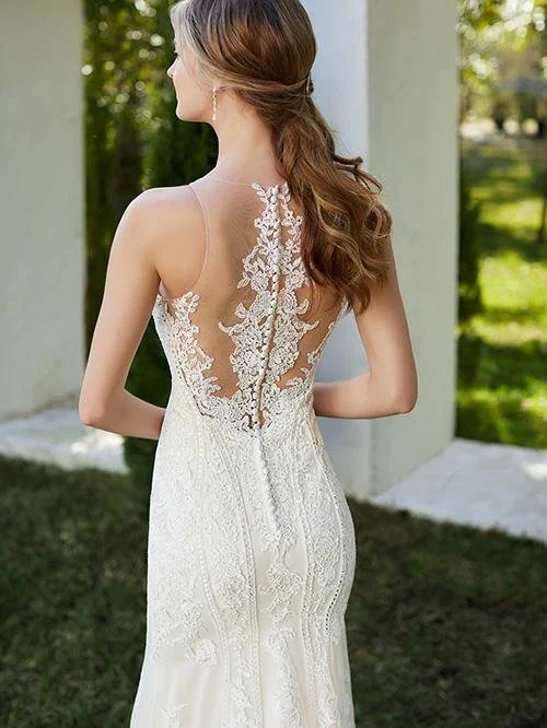 Woman in a white wedding dress with lace details, standing outdoors with greenery in the background.