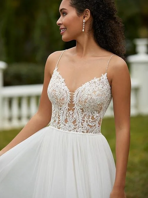 A woman in a white wedding dress outdoors, with lace bodice and thin straps, smiling and looking to her left.