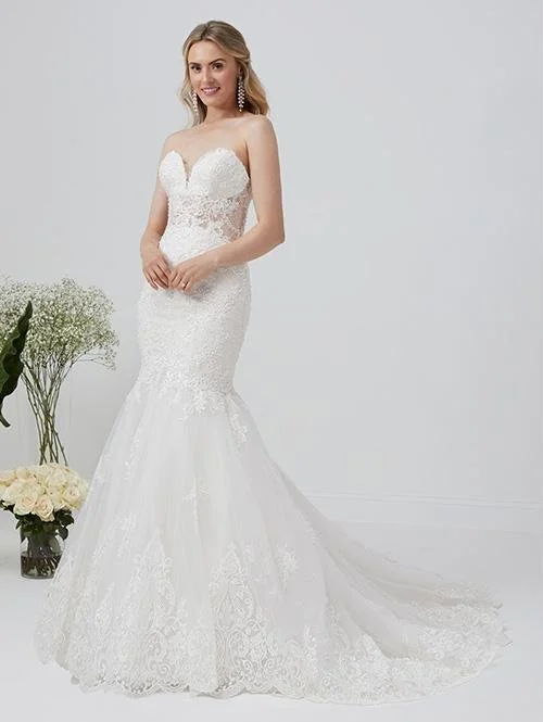 Brunette woman in a white lace wedding gown with a strapless sweetheart neckline, standing in front of a white background, near a bouquet of white flowers and greenery.