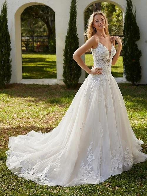 A woman in a white bridal gown standing outdoors in front of a white archway with tall trees in the background.