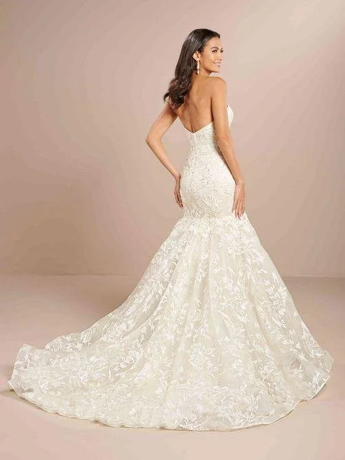 A woman wearing a white wedding gown with a fitted bodice and a full skirt, standing sideways with her hand on her hip, against a plain beige background.