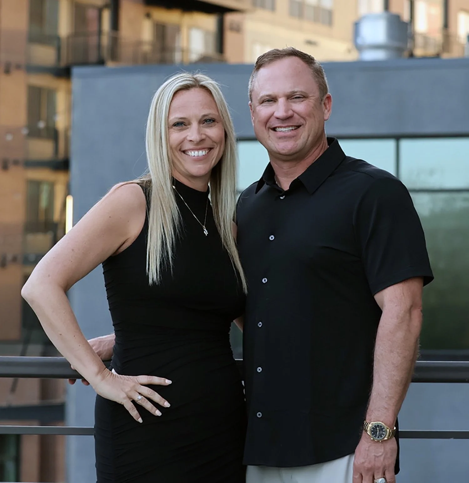 A smiling woman with long blonde hair in a black sleeveless dress, standing next to a smiling man with short hair in a black shirt, outdoors on a balcony with city buildings in the background.
