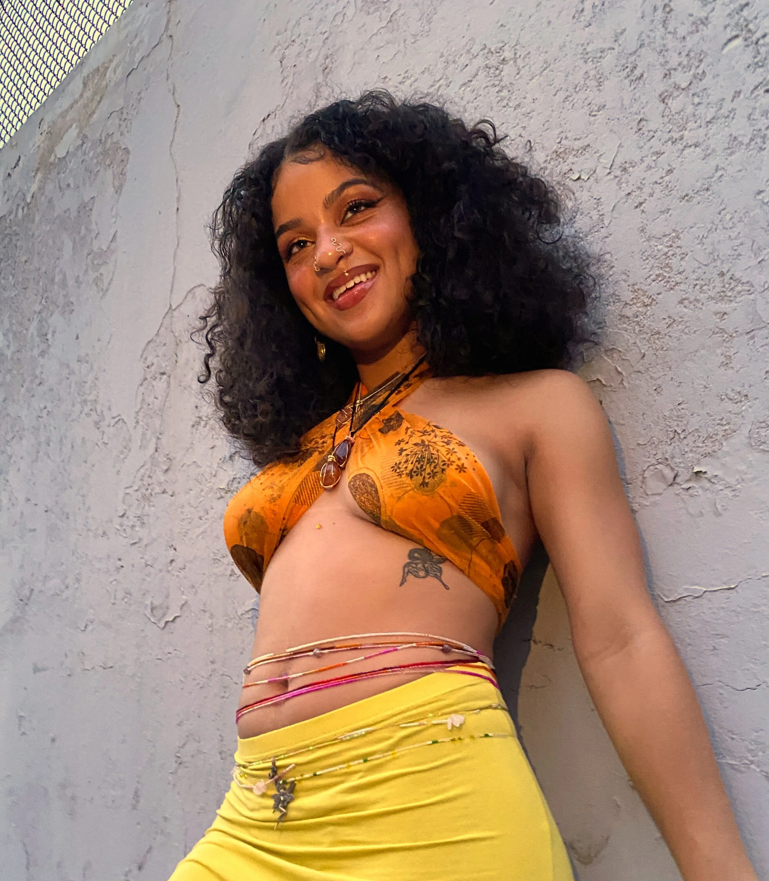 Smiling woman with curly hair, wearing a colorful halter top, yellow pants, and jewelry, leaning against a textured light gray wall.