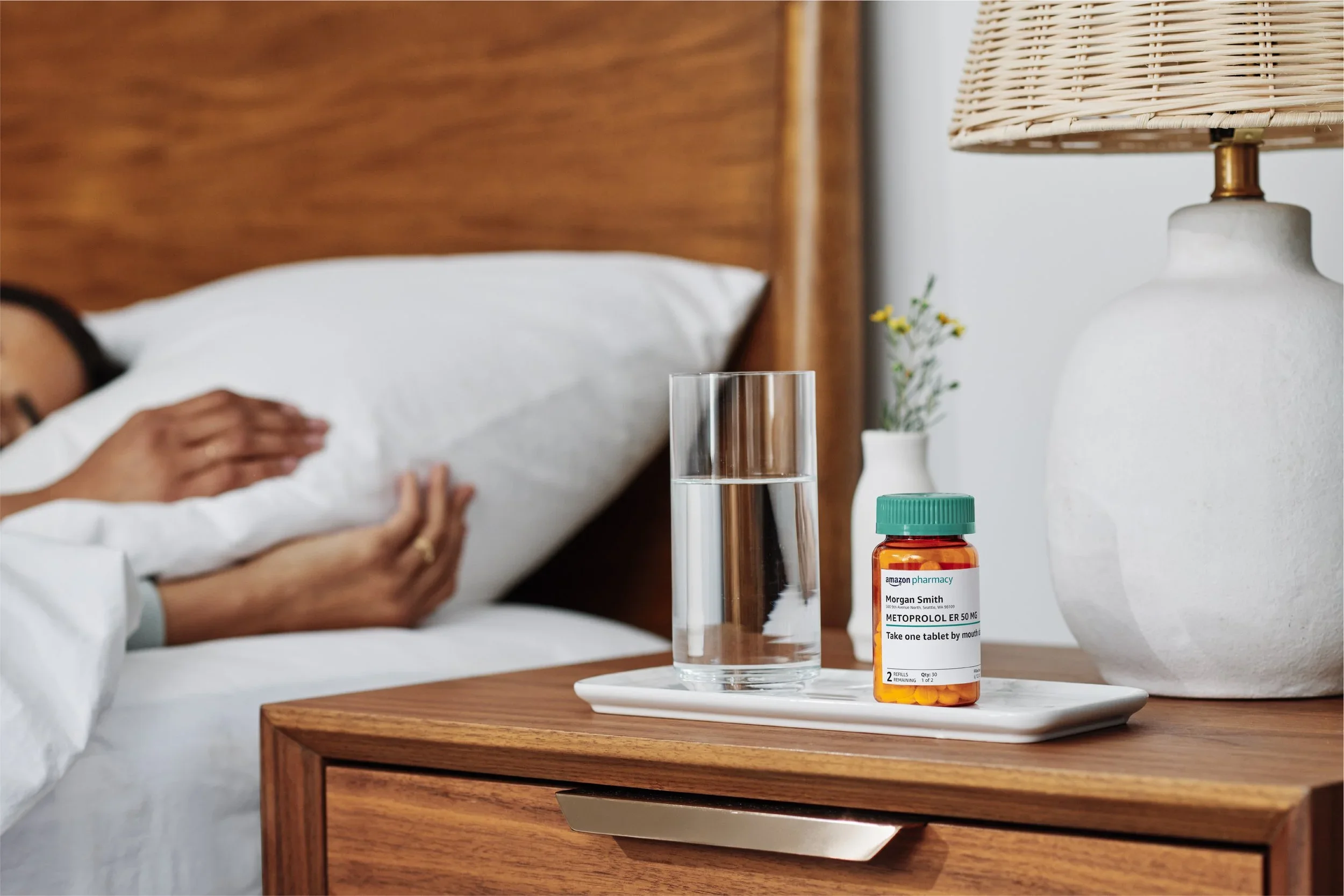 A woman lying in bed with her hand resting on her stomach, next to a wooden nightstand with a glass of water, a bottle of medication labeled 'Metoprolol ER 50 MG,' and a small white vase with a plant. The background includes a wooden headboard and a beige lamp with a woven shade.