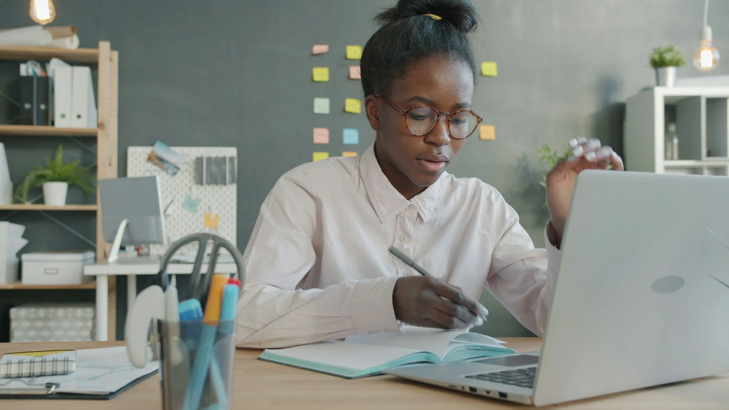 A student works at a desk with their laptop. there are sticky notes on the wall behind her.
