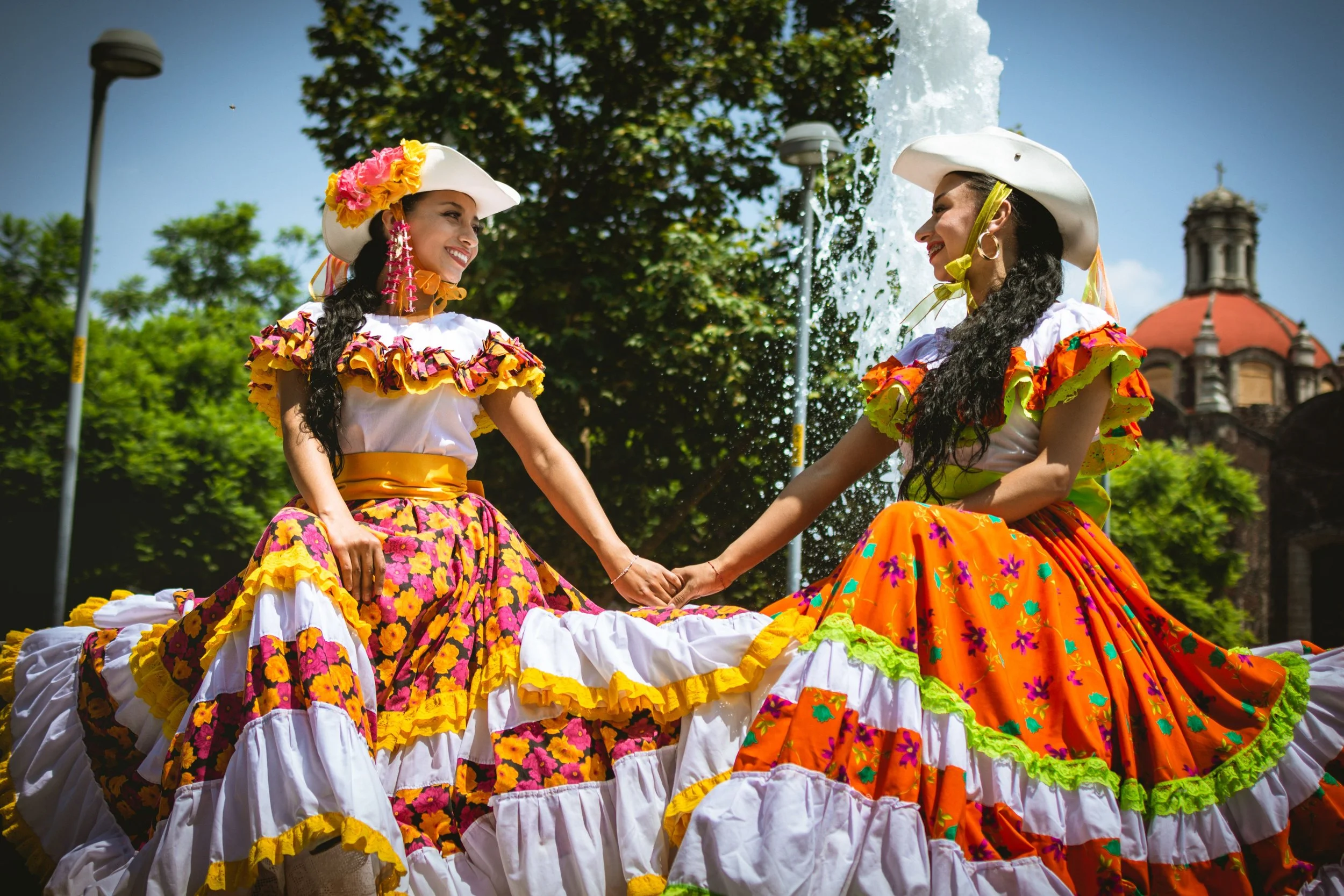 Two women in colorful, traditional Mexican dresses smile at each other holding hands