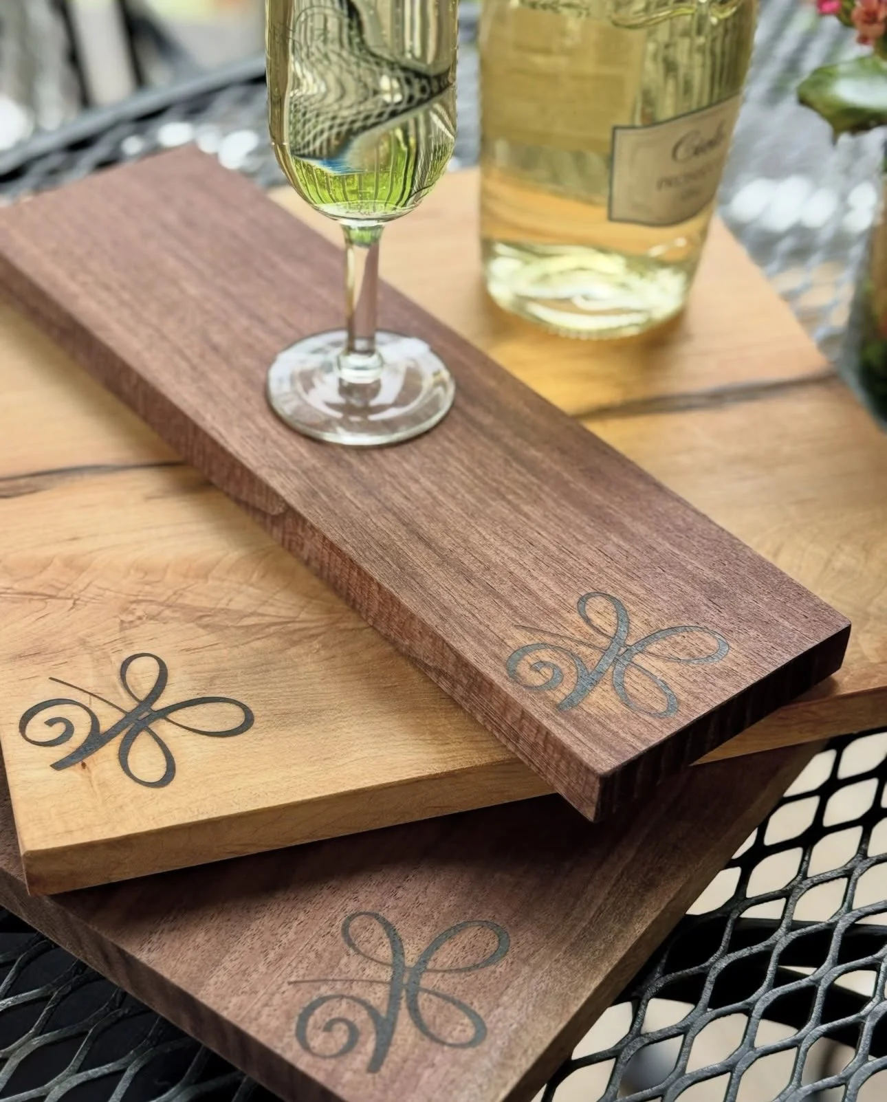 Three wooden drink trays with Fleur-de-lis designs, one with a glass of white wine, on a metal table.