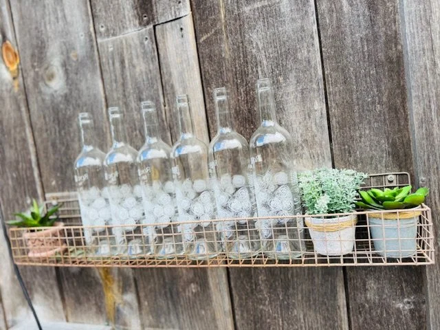 Six empty clear glass bottles placed in a wire basket on a wooden surface, accompanied by potted plants.
