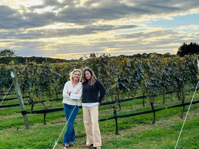 Two women standing in a vineyard during sunset, with grapevines behind them and a cloudy sky above.