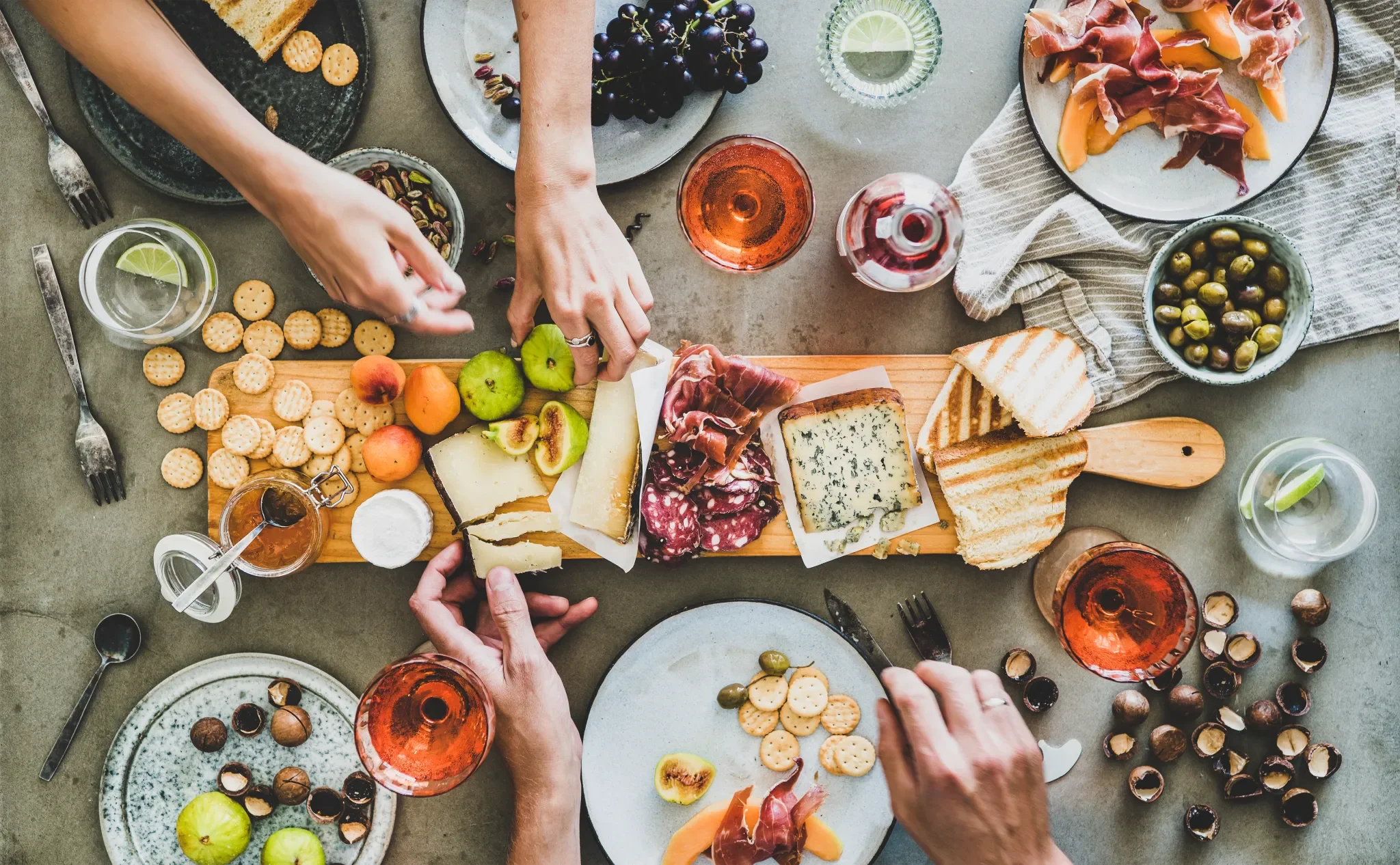 Top view of a cheese and charcuterie board with fruits, crackers, wine glasses, and people’s hands reaching for food at a social gathering.