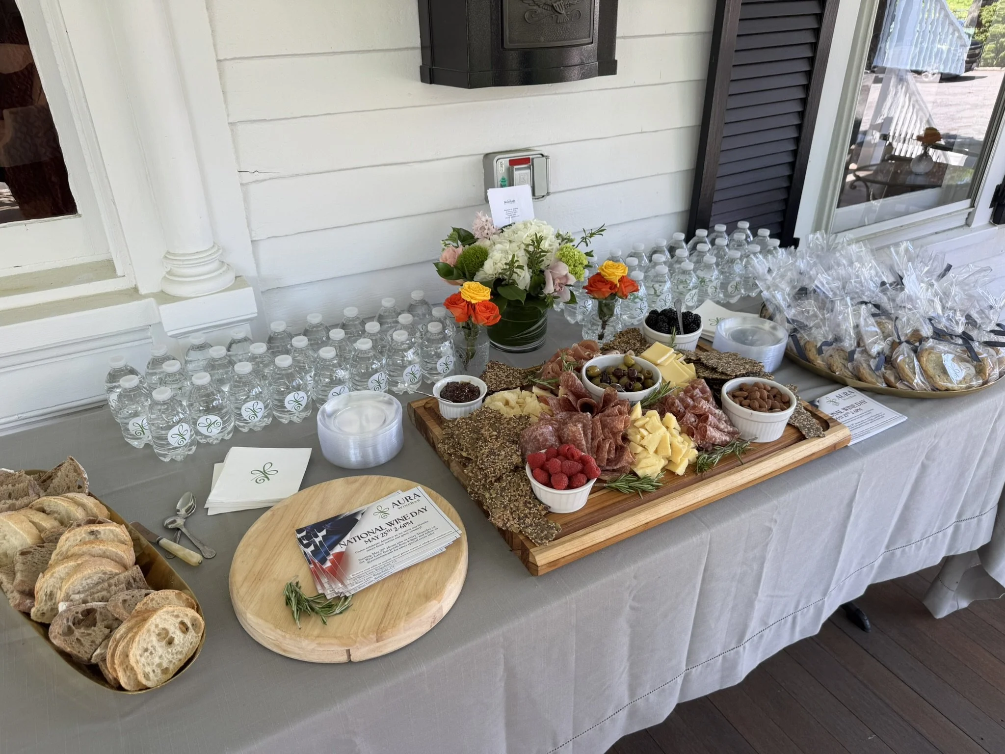 A cheese and charcuterie platter with bread, fruit, and snacks, alongside bottled water and flowers, set on a table for a special event.