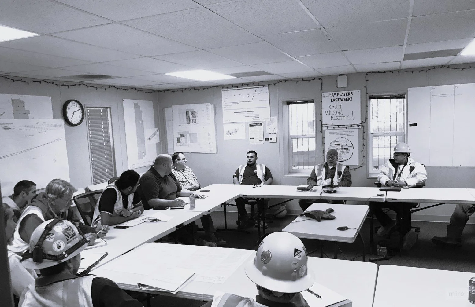 Reunión en una sala de trabajo con varias personas, algunos con casco de seguridad, en un ambiente laboral.