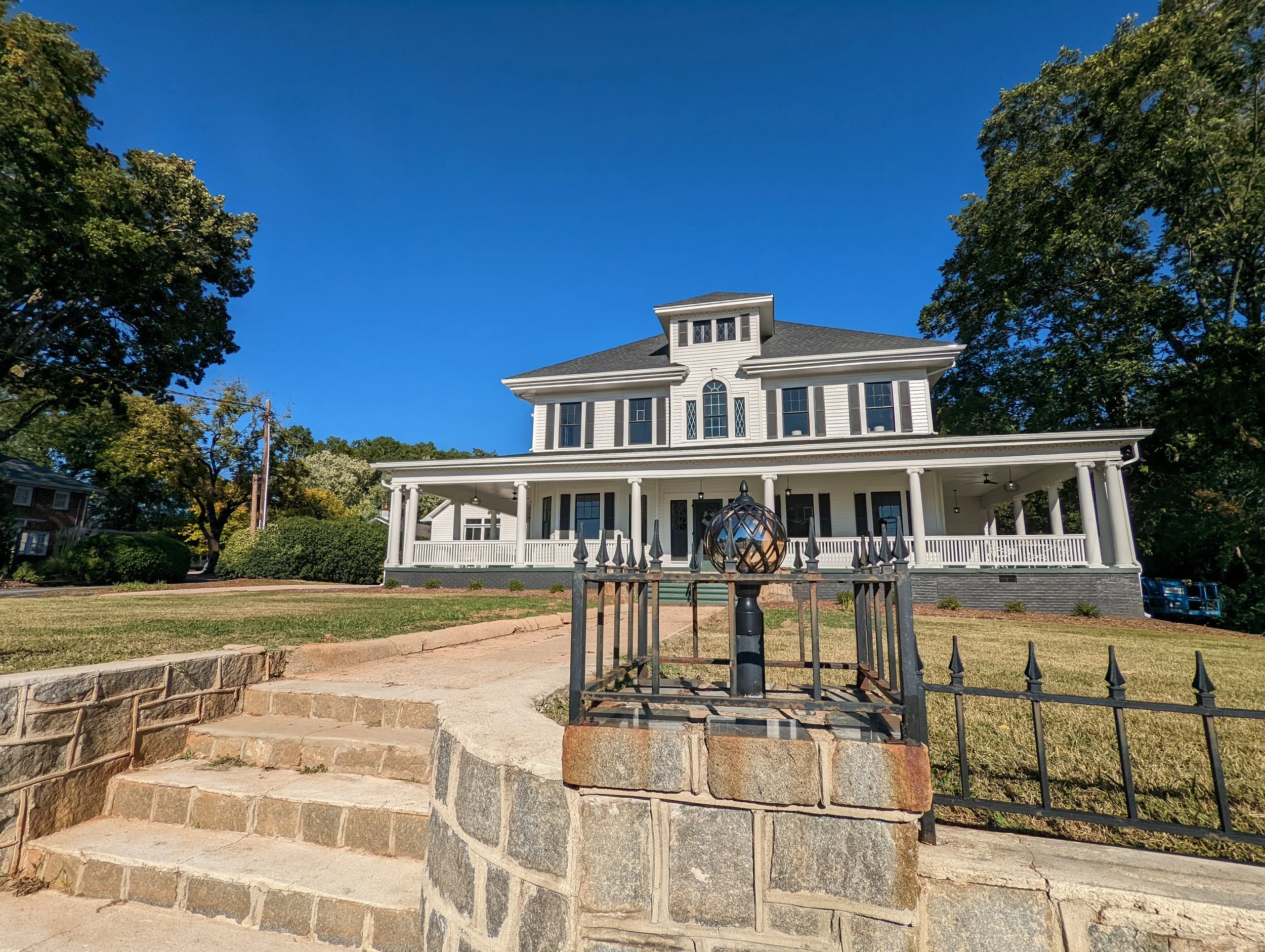 Restored Historic Hampton House in Pickens, SC, featuring a grand two-story exterior, wraparound porch, white siding, stone entrance steps, and decorative wrought-iron fencing.