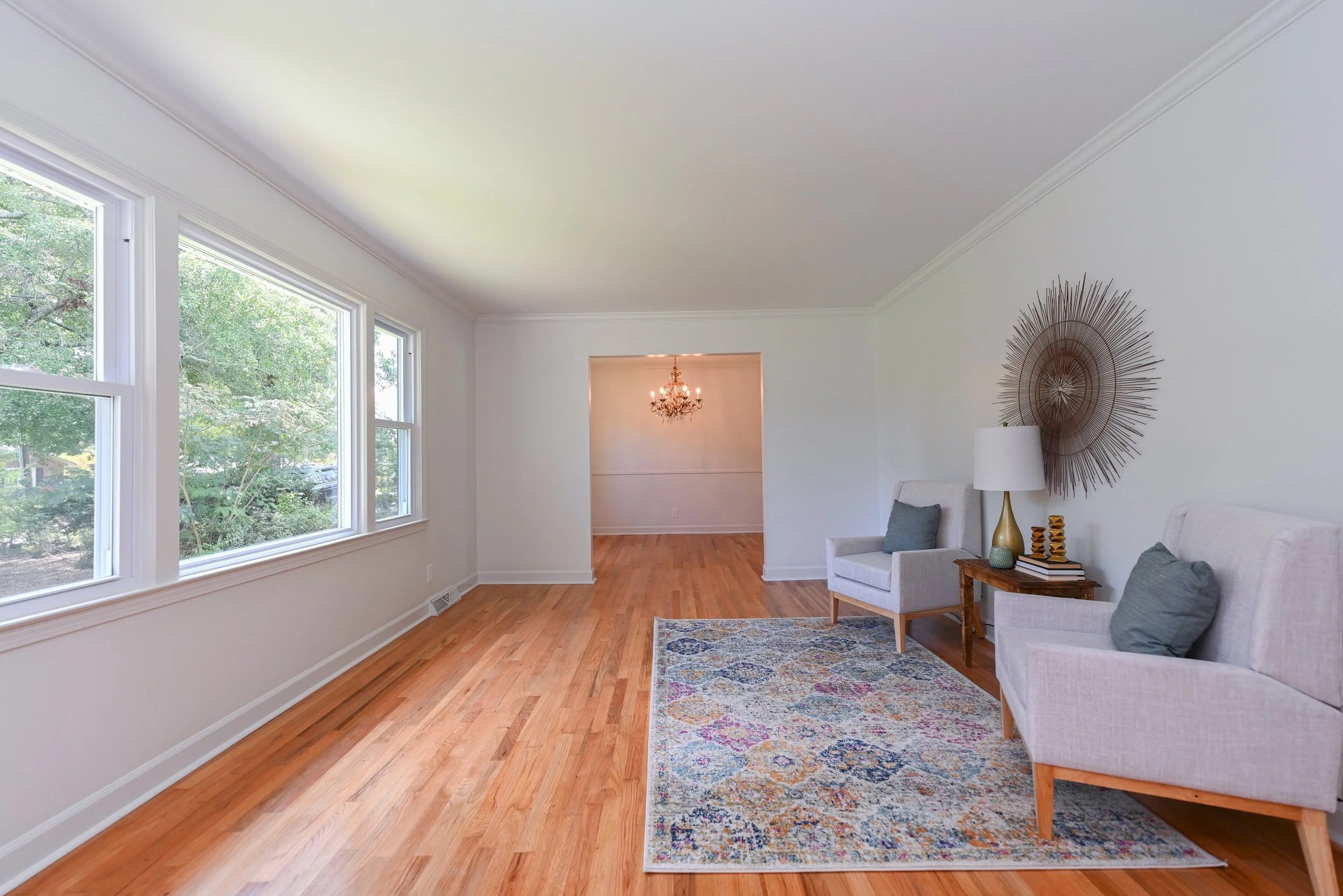 Renovated living room at 33 Oriole Street in Greenville, SC, featuring hardwood floors, large windows overlooking mature trees, white walls, and an open transition to the adjoining dining area.