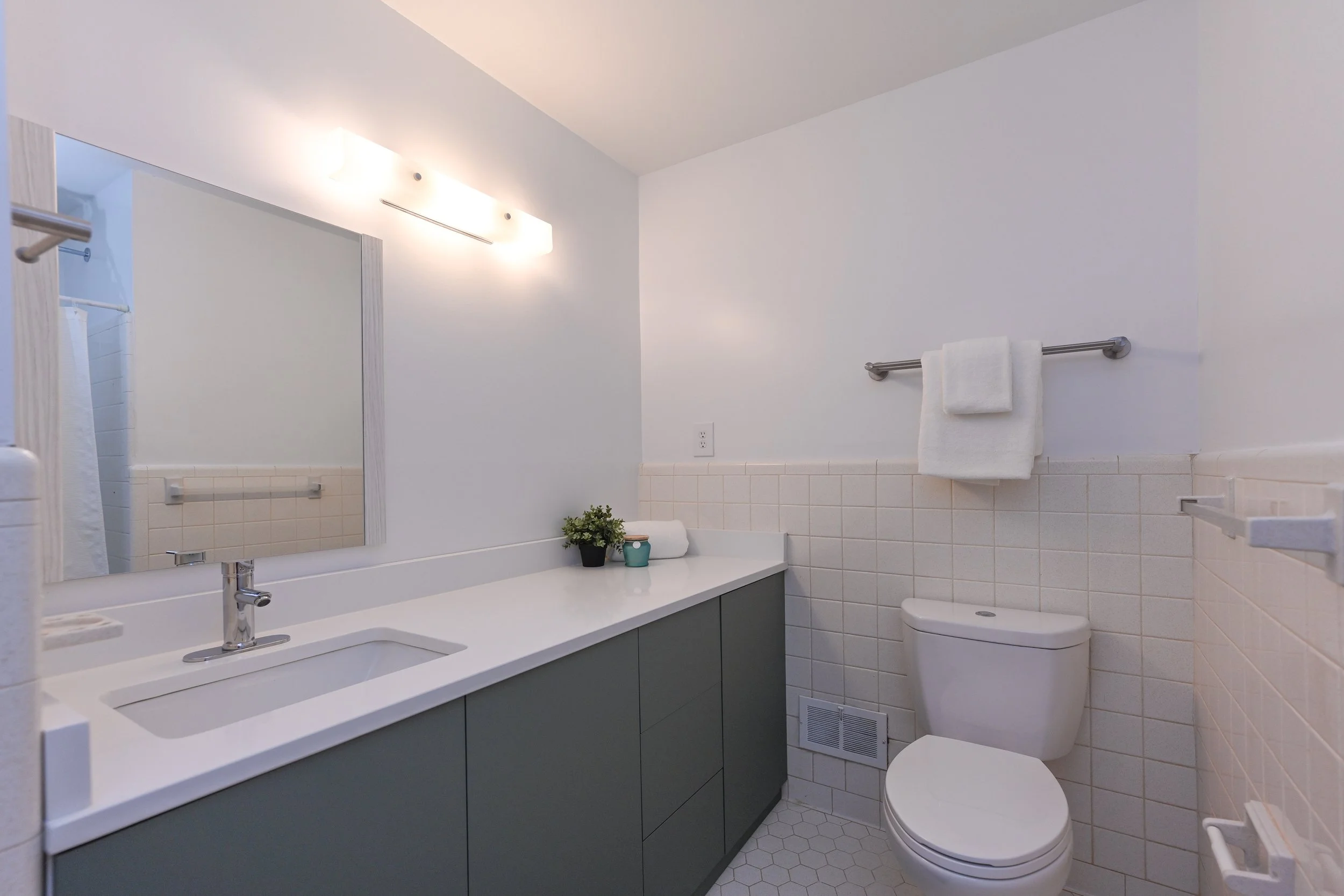 Renovated bathroom at 33 Oriole Street in Greenville, SC, featuring a modern gray vanity, white countertop, chrome fixtures, white tiled walls, and hexagonal floor tiles.
