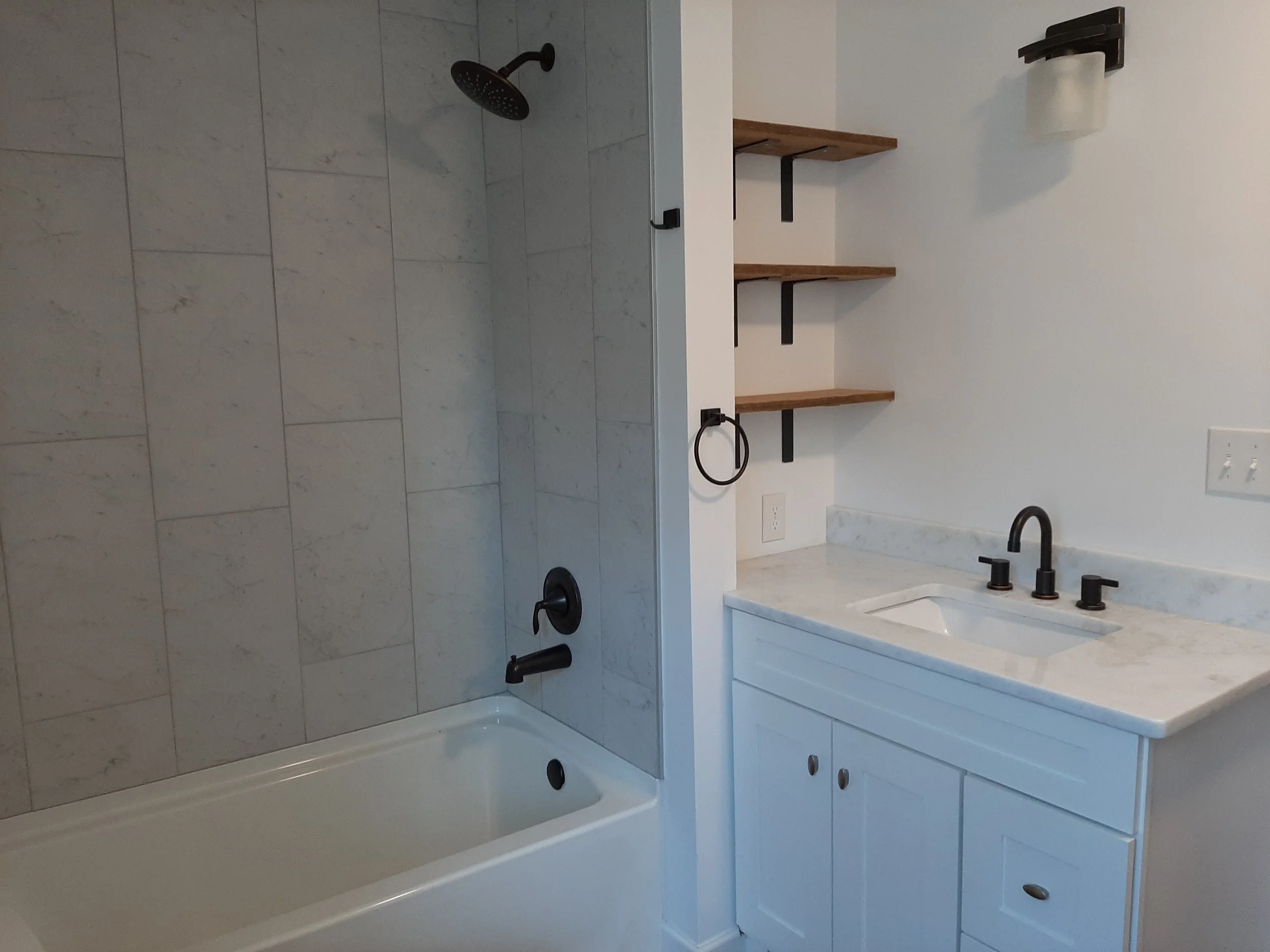 Renovated guest bathroom at 104 Medallion Lane in Lyman, SC, featuring marble-look shower tiles, a white vanity, black fixtures, and open wood shelving.