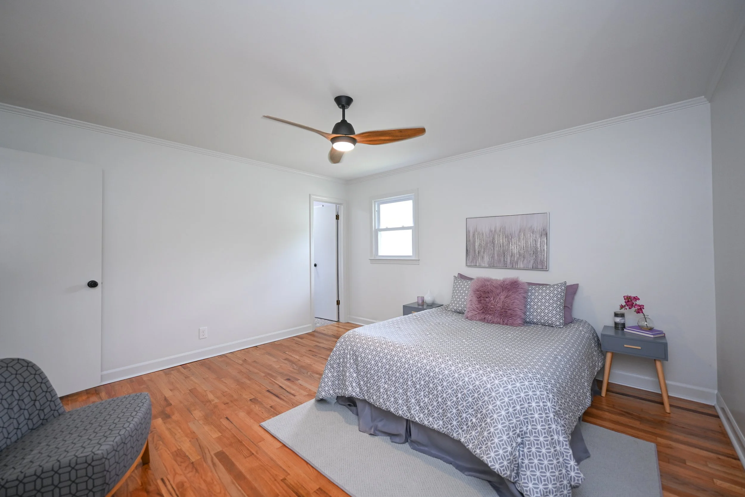 Renovated bedroom at 33 Oriole Street in Greenville, SC, featuring warm hardwood flooring, white walls, a modern ceiling fan, and natural light from a window.