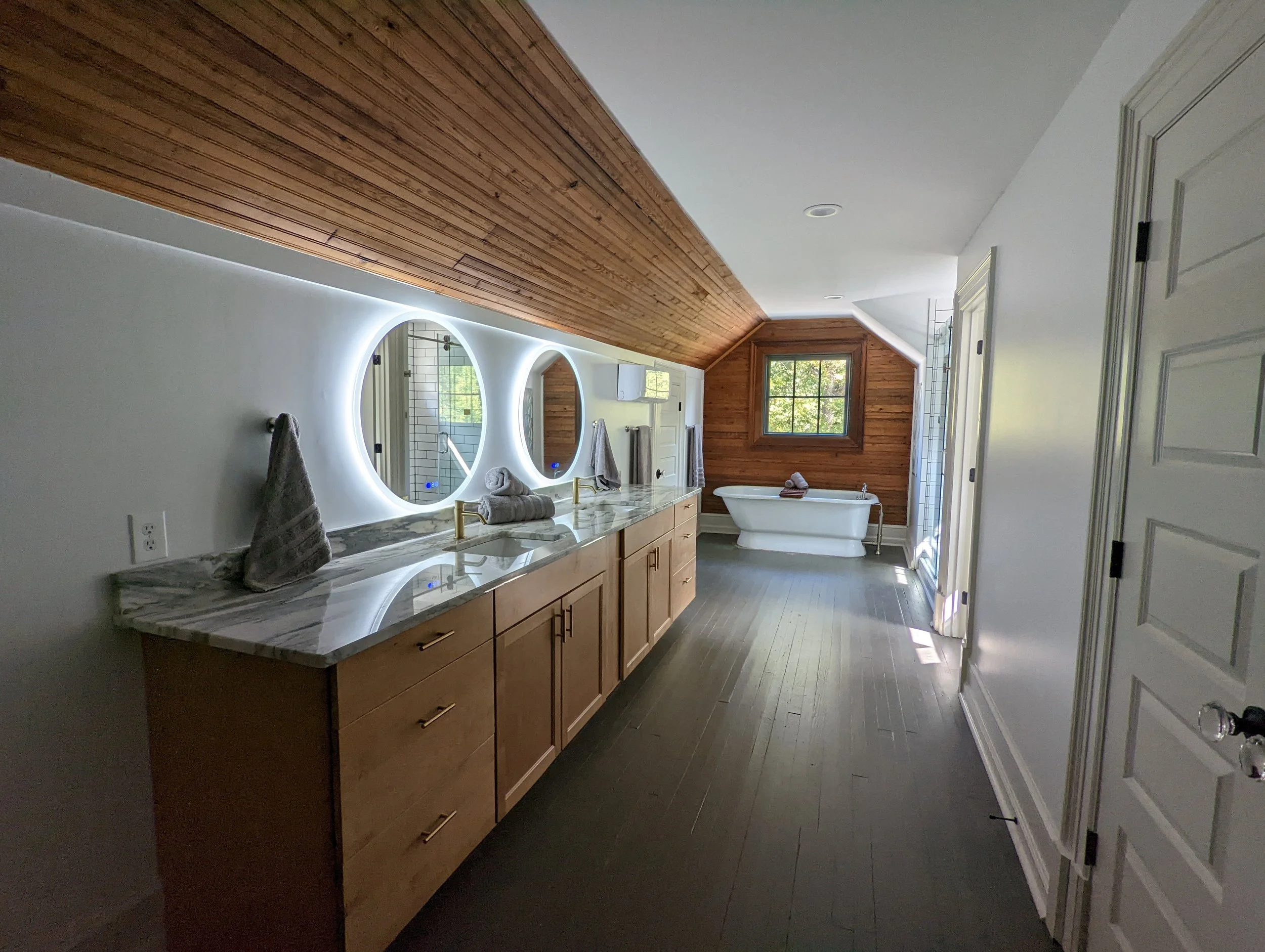 Renovated bathroom at Historic Hampton House 1908 in Pickens, SC, featuring a double vanity, illuminated round mirrors, wood-paneled ceiling, and freestanding bathtub.