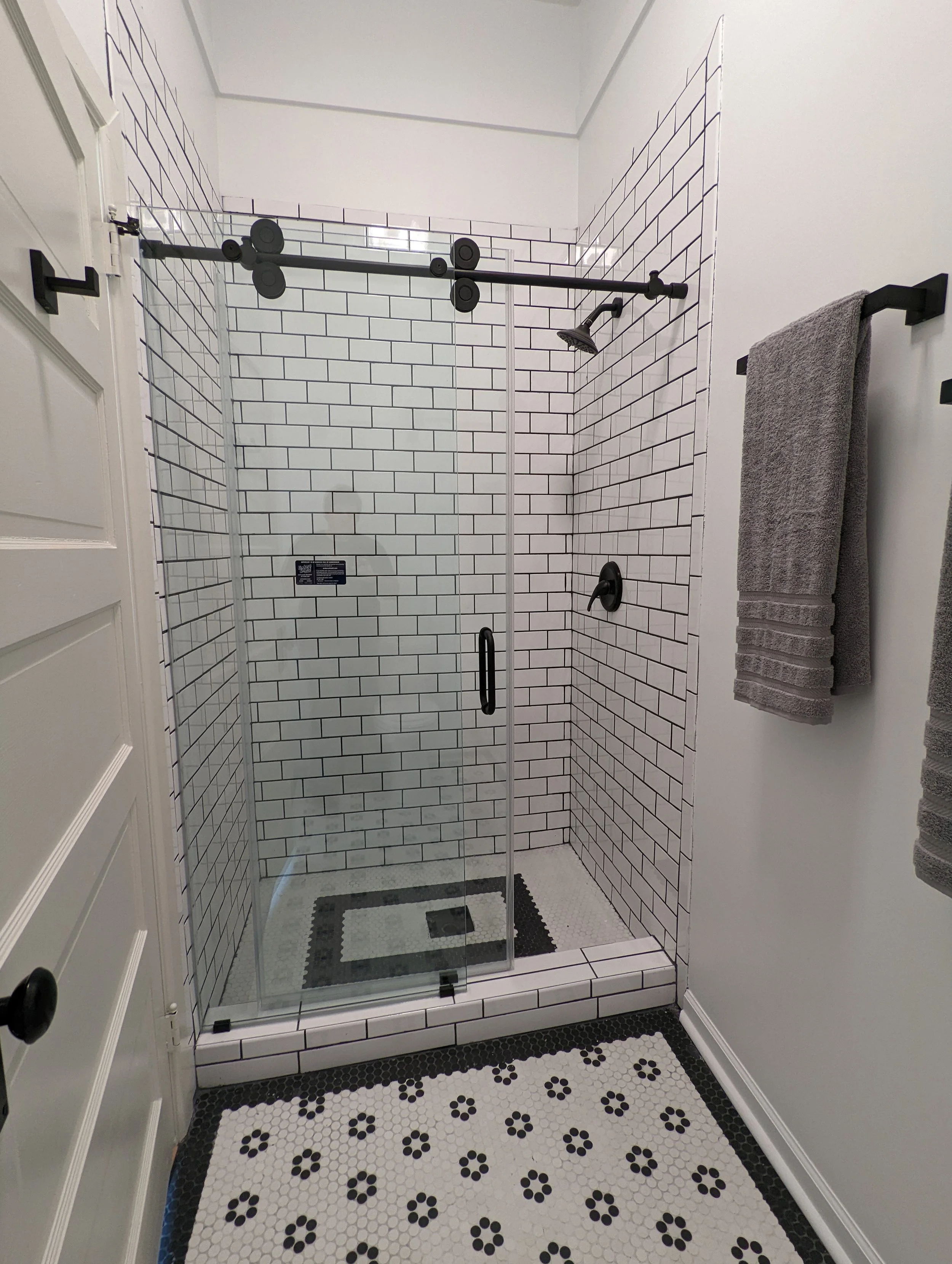 Renovated walk-in shower at Historic Hampton House 1908 in Pickens, SC, featuring white subway tile, black fixtures, a glass enclosure, and decorative black-and-white mosaic floor tile.