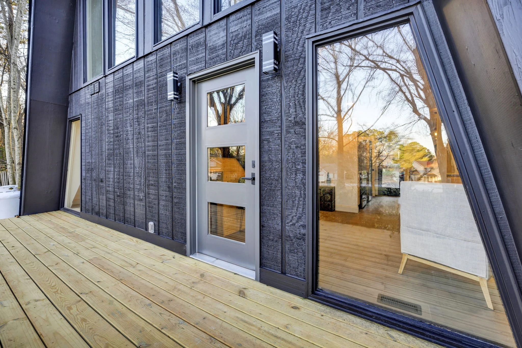 Renovated home at 112 Wayland Drive in Duncan, SC, featuring dark board-and-batten siding, a modern glass-panel entry door, floor-to-ceiling windows, and a natural wood deck.