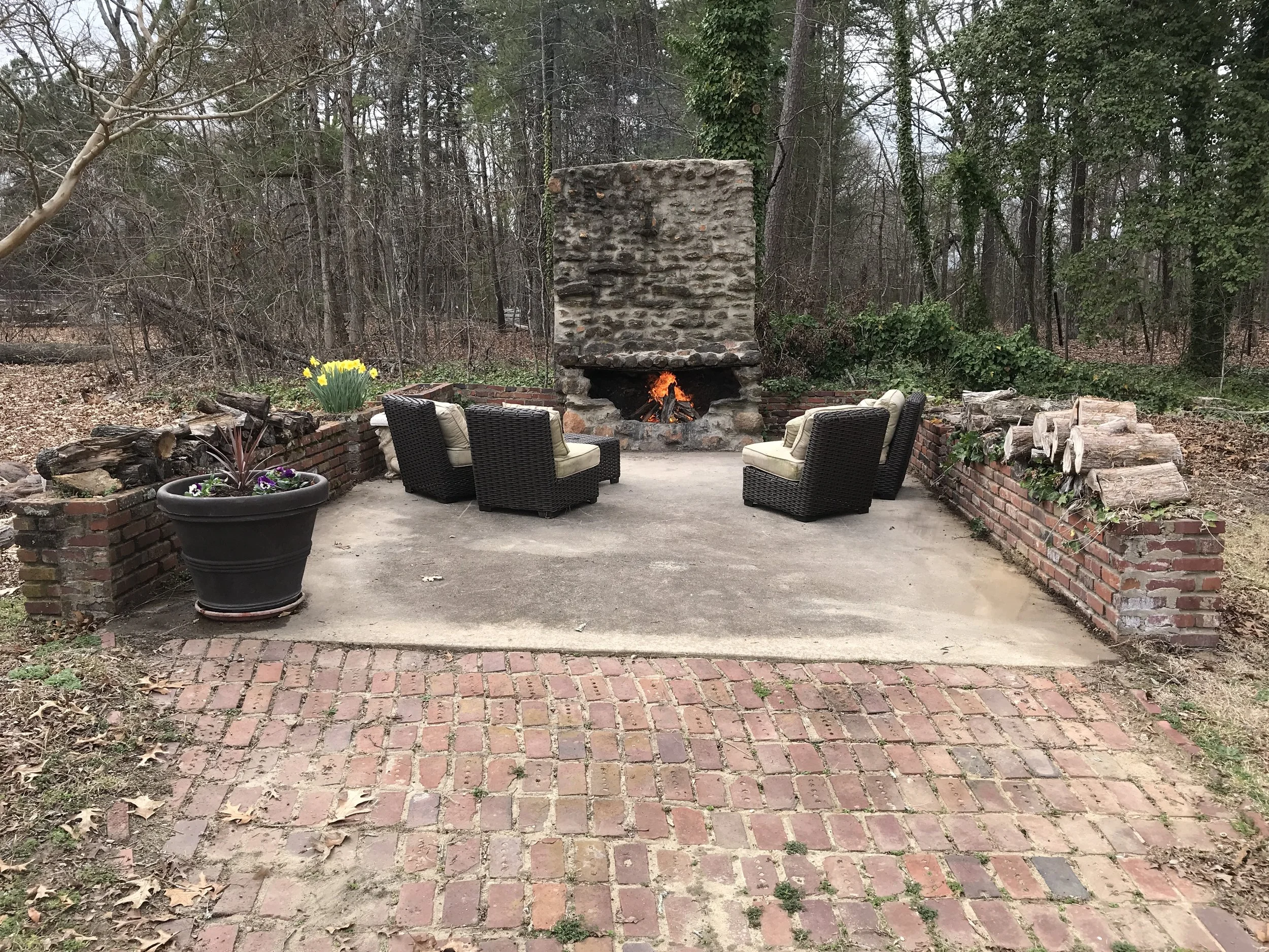 Outdoor entertaining patio at 104 Medallion Lane in Lyman, SC, featuring a rustic stone wood-burning fireplace, brick paving, and seating surrounded by mature trees.