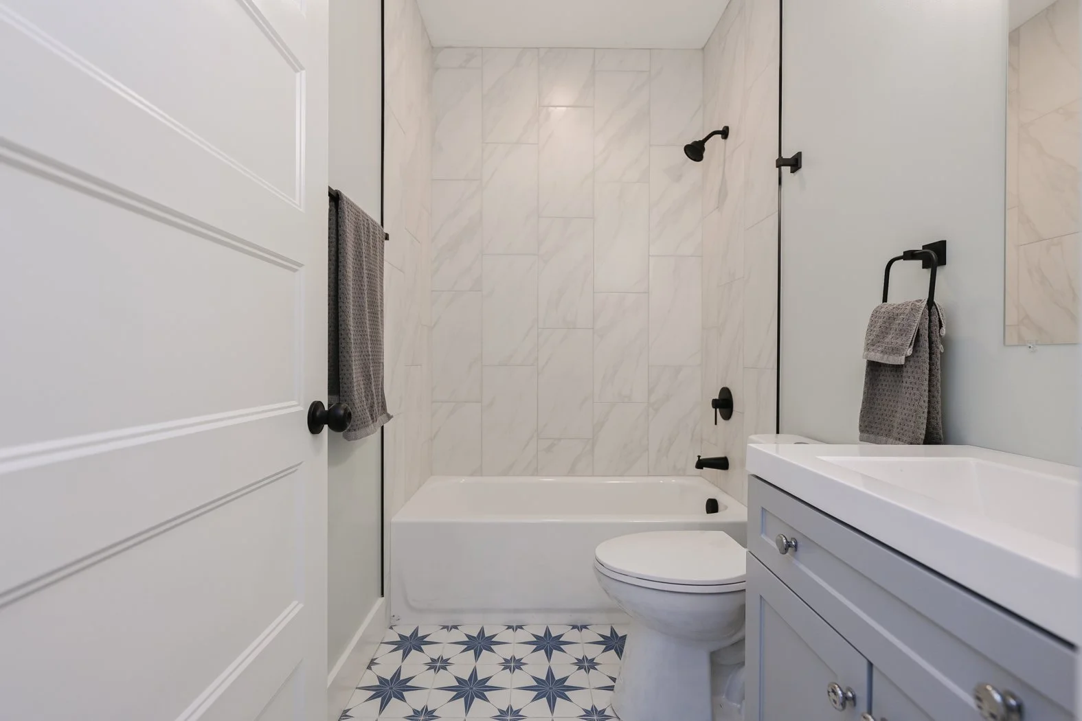 Renovated guest bathroom at 1334 Jameson Road in Easley, SC, featuring marble-look bathtub tiles, matte black fixtures, a gray vanity, and blue-and-white patterned floor tiles.