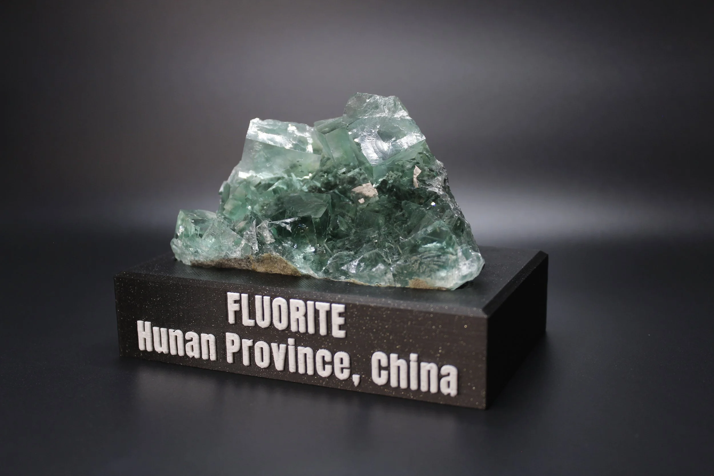 A mineral specimen of fluorite from Hunan Province, China, displayed on a black stand with white lettering.