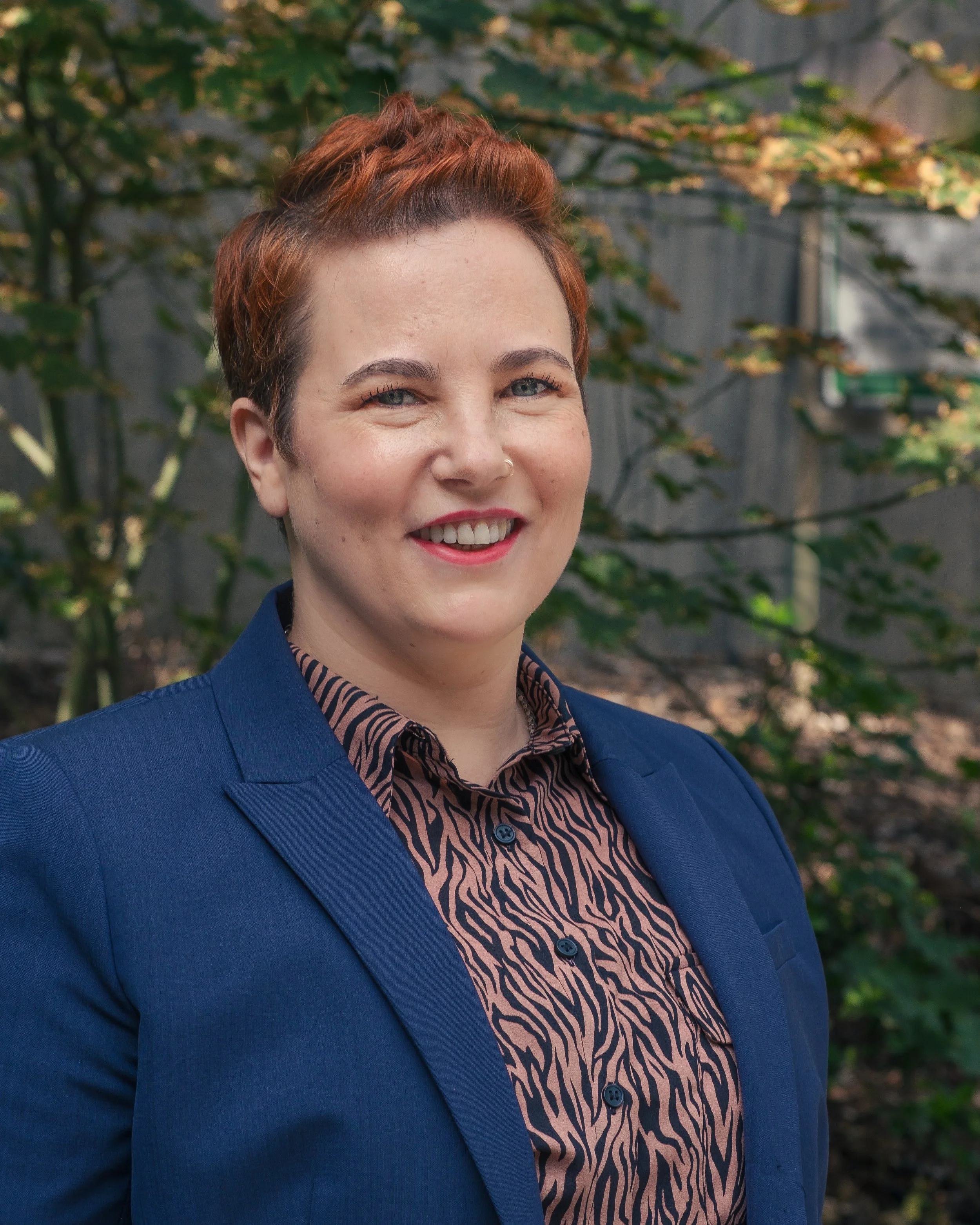 Alexa Lachman wearing a navy blazer and a brown and black patterned shirt, with trees and greenery in the background.