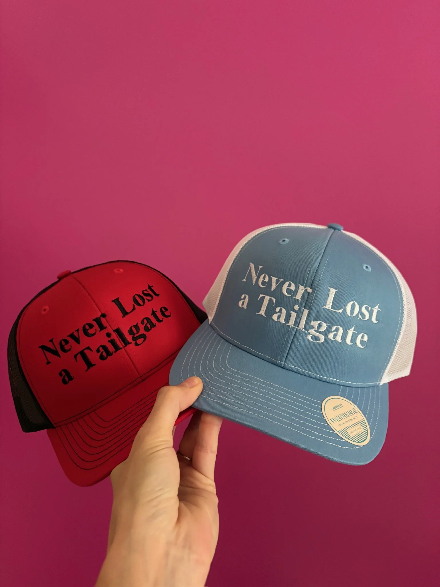Two trucker hats, one red and one blue, being held up against a pink background. The red hat has the words 'Never Lost a Tailgate' in black text, while the blue hat has the same phrase in white text.