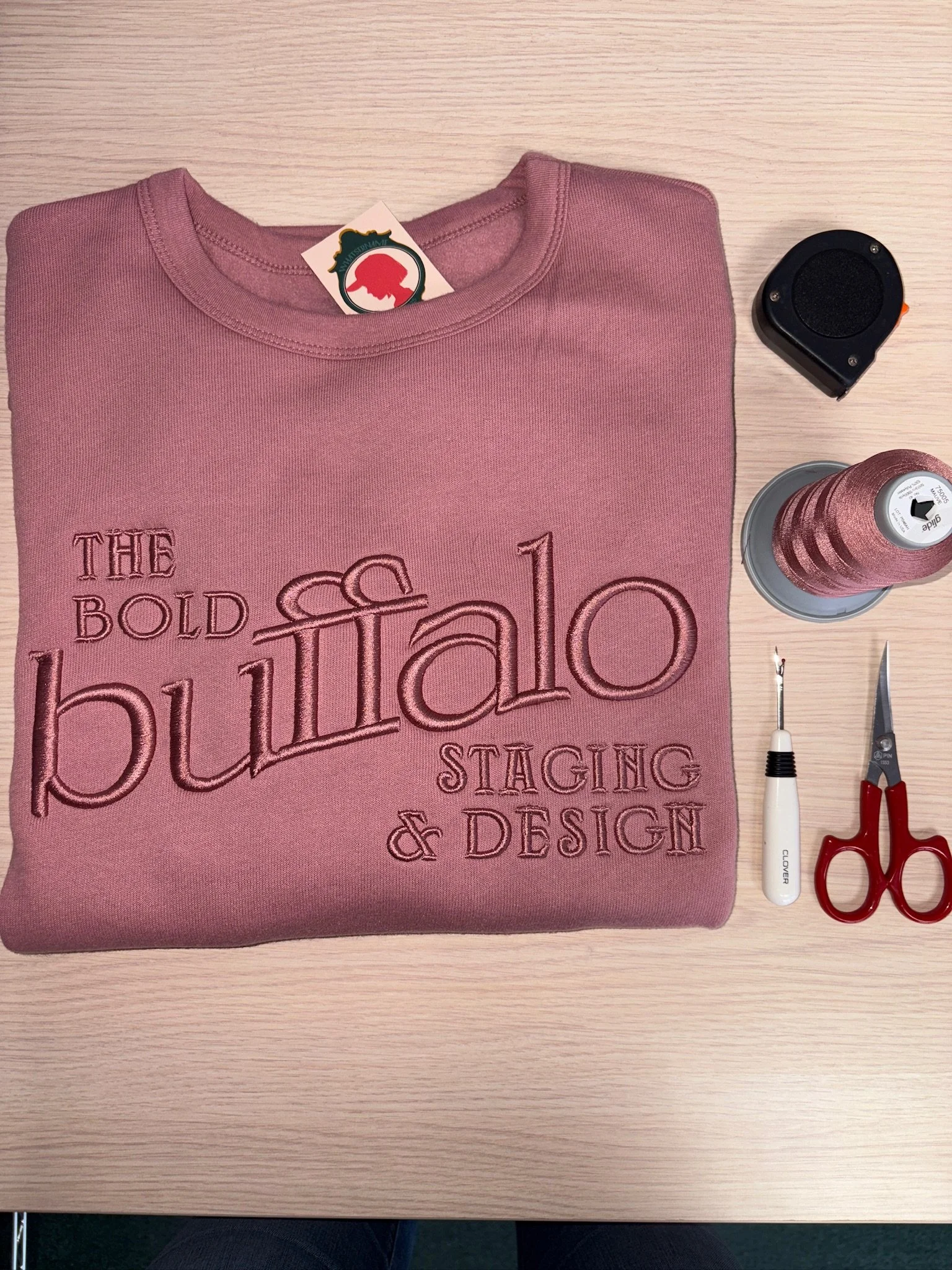A pink sweatshirt with embroidered local small business branding  'The Bold Buffalo Staging & Design' is folded on a wooden table with sewing tools including a measuring tape, a bobbin of pink thread, a seam ripper, and small scissors.