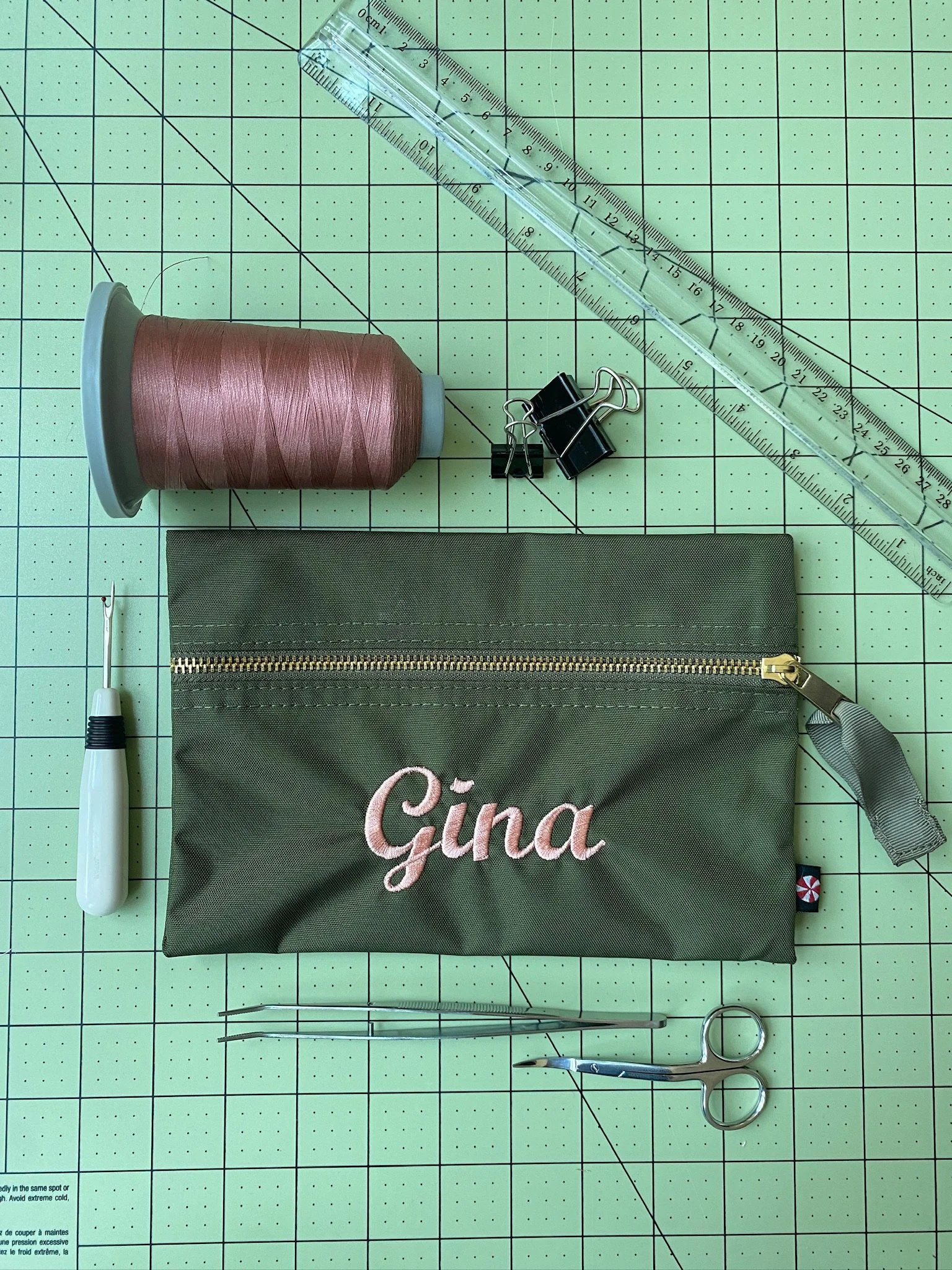 Flat lay of sewing supplies on a cutting mat: pink thread on a spool, black binder clips, a clear acrylic ruler, a seam ripper, small scissors, and a green zippered pouch embroidered with the name 'Gina'.