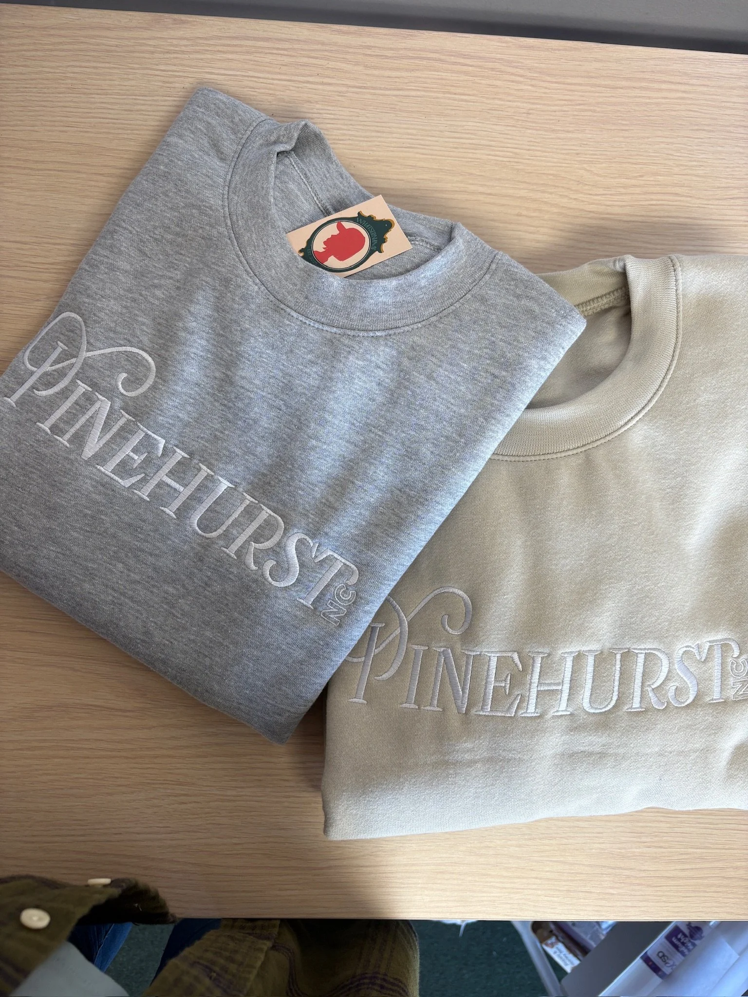Two folded sweatshirts, one gray and one cream, both with embroidered text. The gray sweatshirt has the word 'PINEHURST' and the cream sweatshirt also has the word 'PINEHURST,' both in large embroidered letters.
