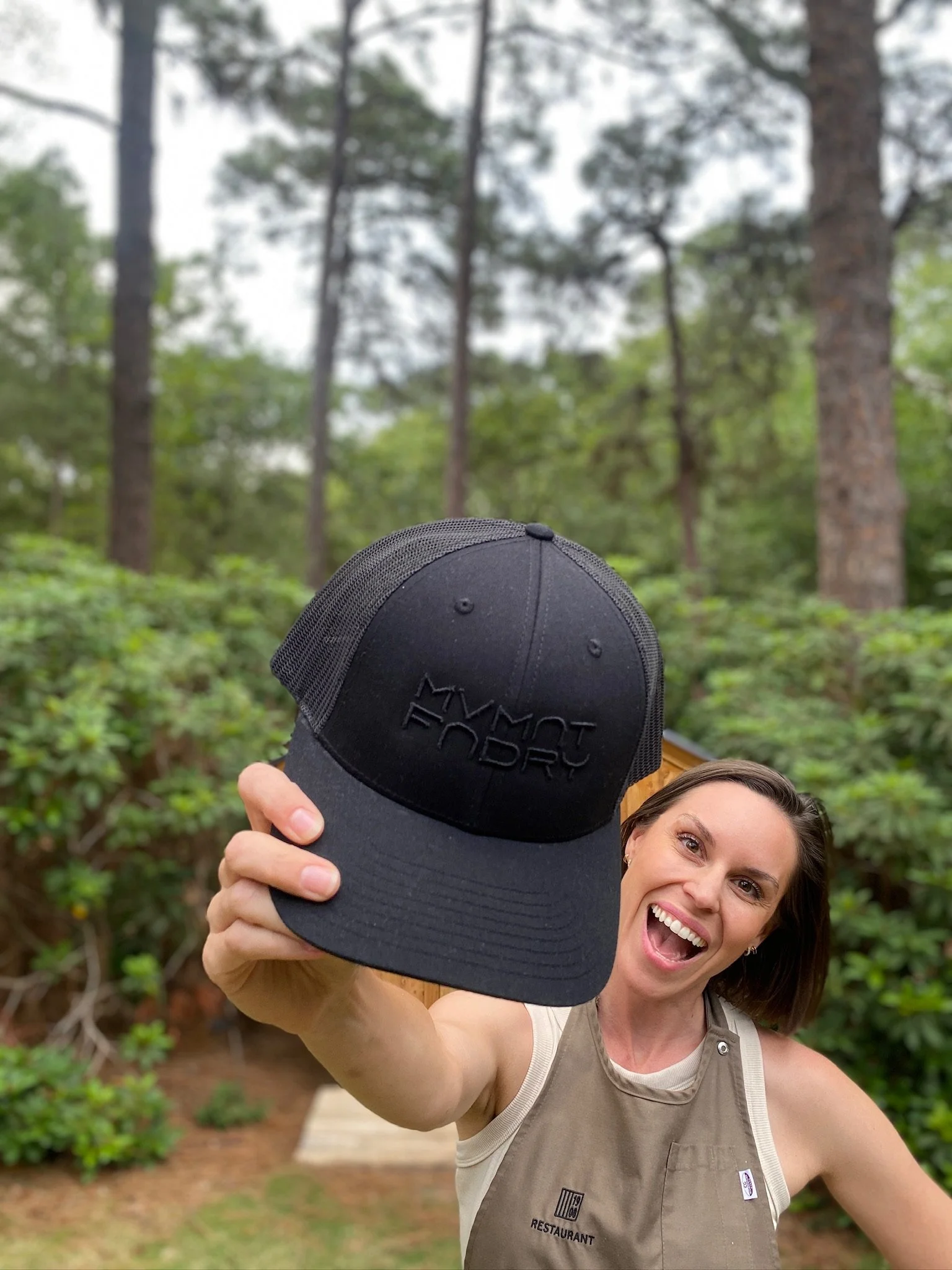 A woman smiling and holding a black cap labeled "Mynot Factory" in front of her in an outdoor forest setting.