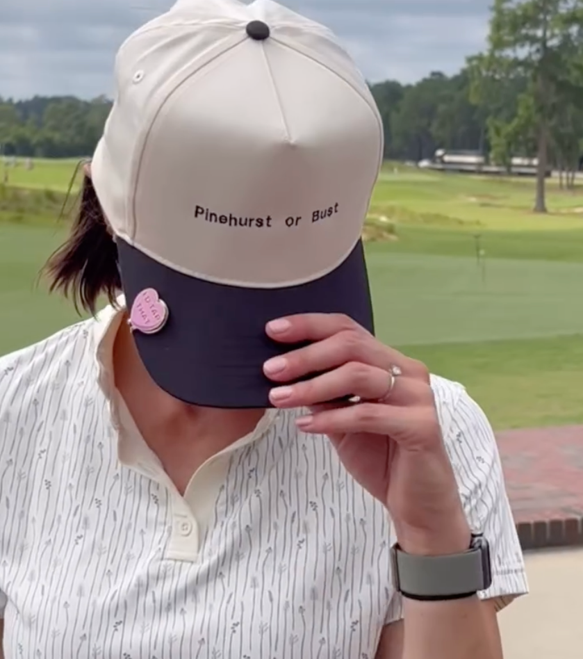 A woman wearing a white and navy cap with the words 'Pinehurst or Bust' embroidered on it, shielding her eyes with her hand, standing outdoors at a golf course.