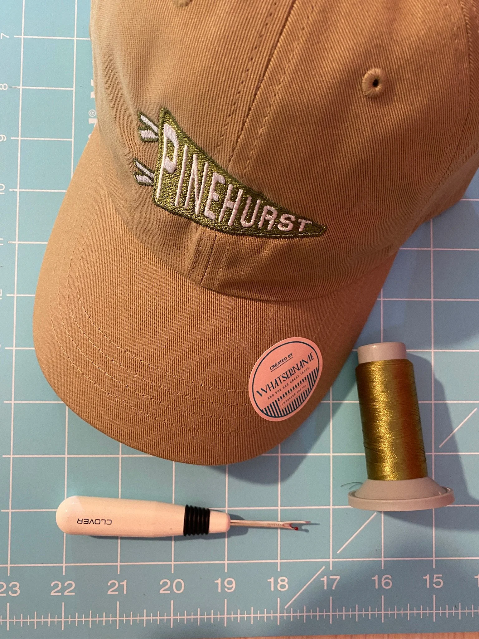 A brown baseball cap with embroidered patch reading 'PINEHURST' in green and white, a white seam ripper with black grip, and a spool of gold thread on a blue grid cutting mat.