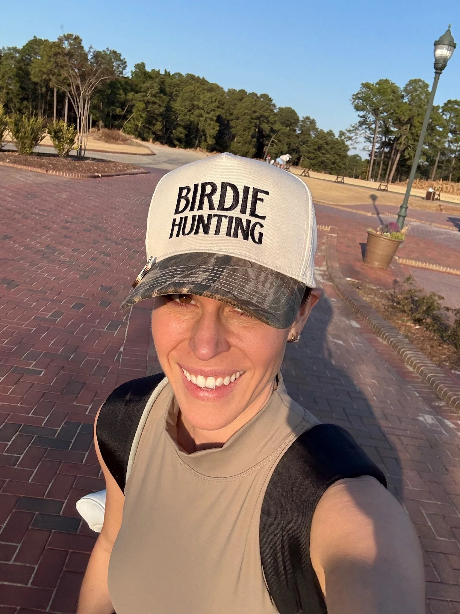 A woman taking a selfie outdoors on a paved path in a park, wearing a cap that says 'Birdie Hunting' and a beige athletic top, with trees and a lamppost in the background during late afternoon sunlight.