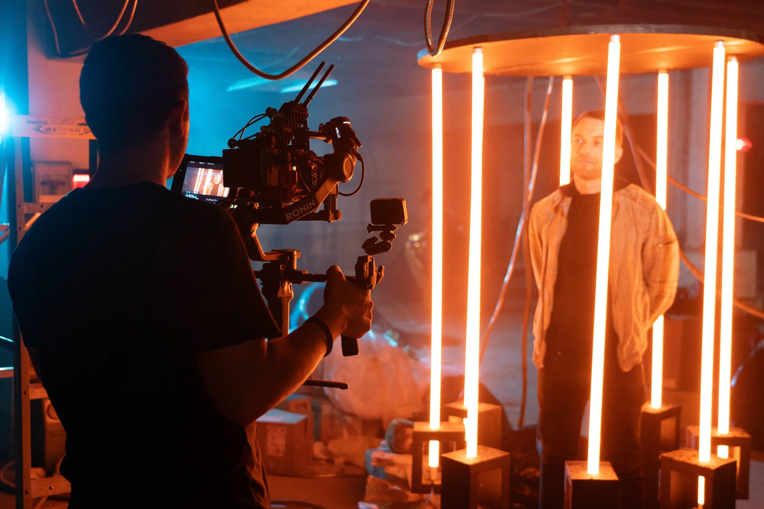 A person filming with a professional camera on a stabilizer captures a man standing behind glowing vertical light tubes in a dimly lit setting.