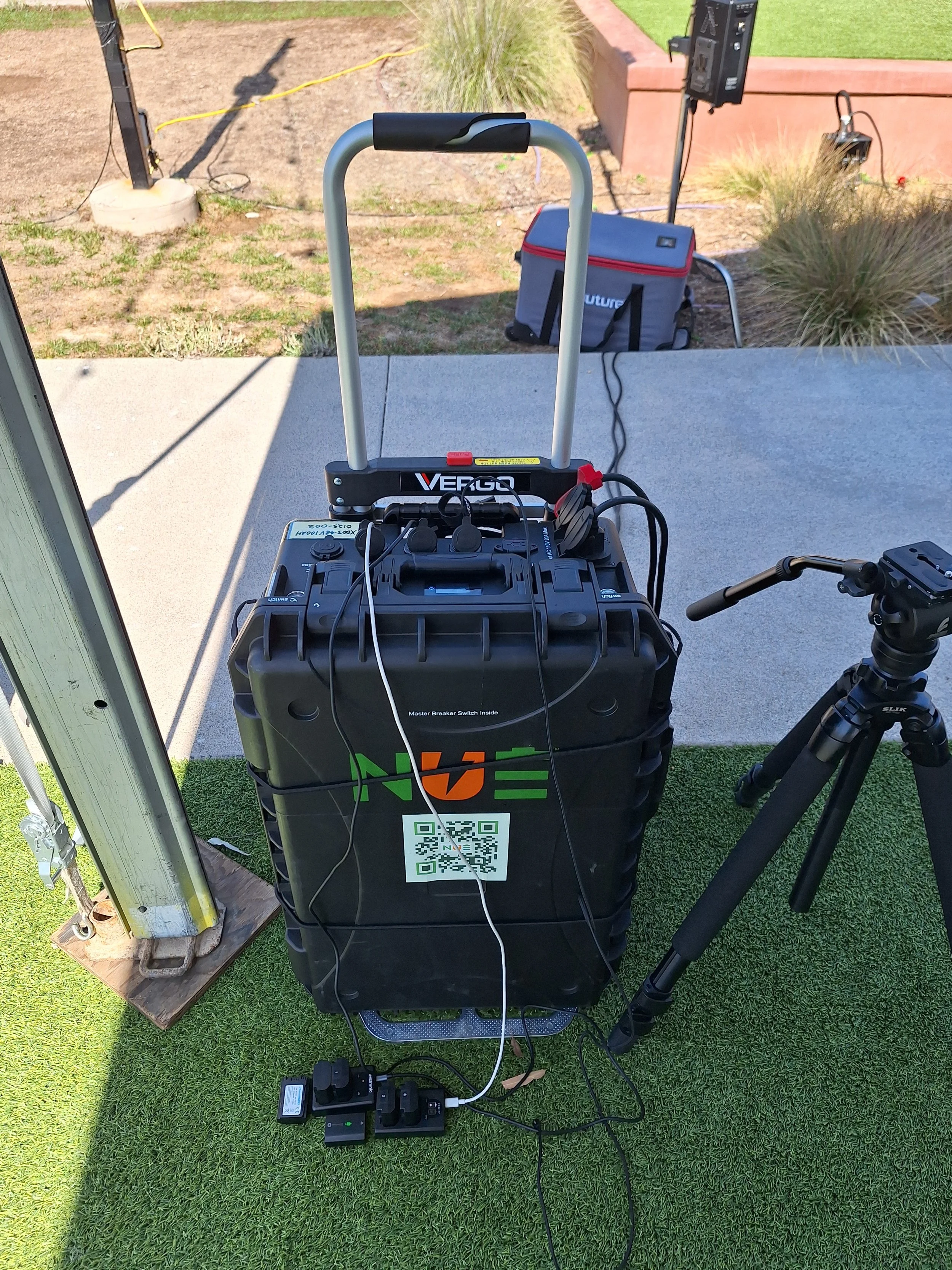 A portable emergency power generator with multiple cables connected, placed on artificial grass outdoors. Behind it is a tripod and a bag, with a background of a patio, plants, and a lighting stand.