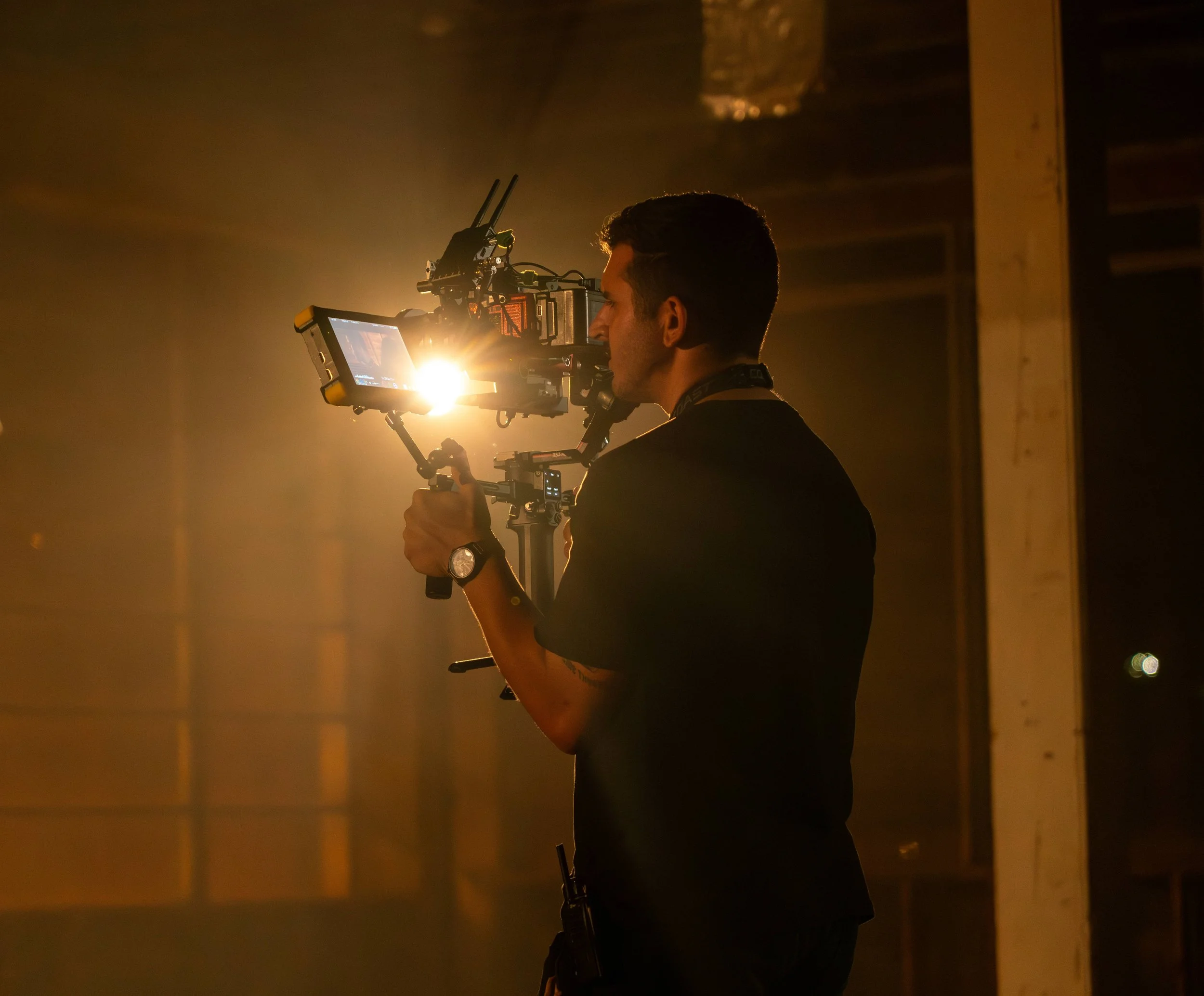 A man operating a professional camera on a film set, illuminated by a bright light, with a dark workshop background.
