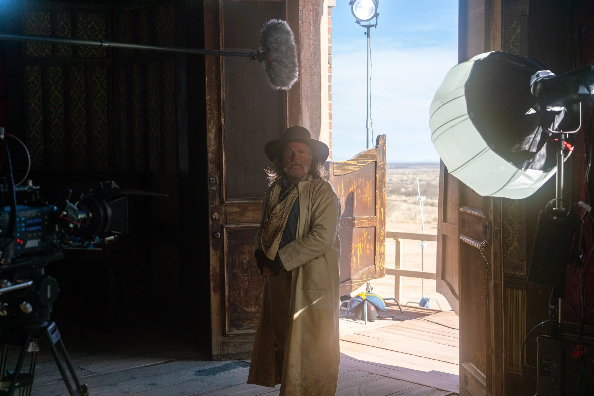A person in period costume standing in a doorway with film equipment and a desert landscape in the background.
