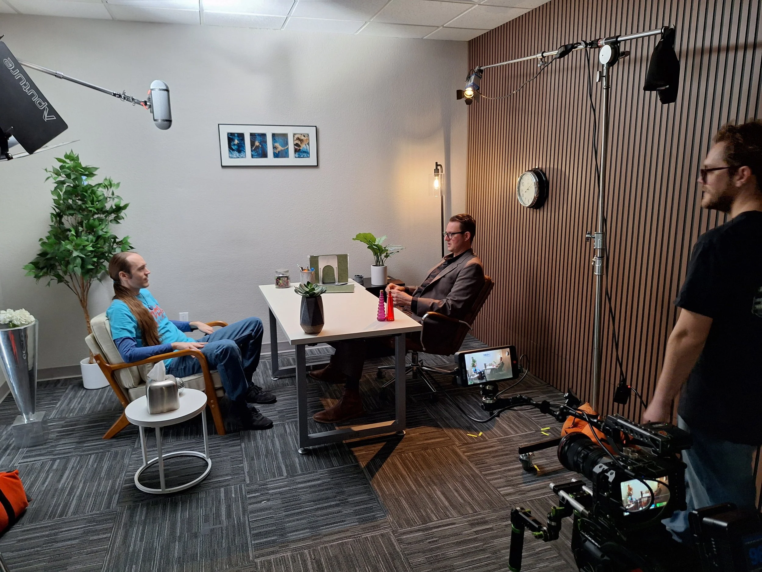 A person with long hair wearing a blue shirt and jeans sits in a chair facing a man in a suit at a desk in a recording studio or interview setting. Two crew members with camera equipment film the scene, with lighting and plants in the background.