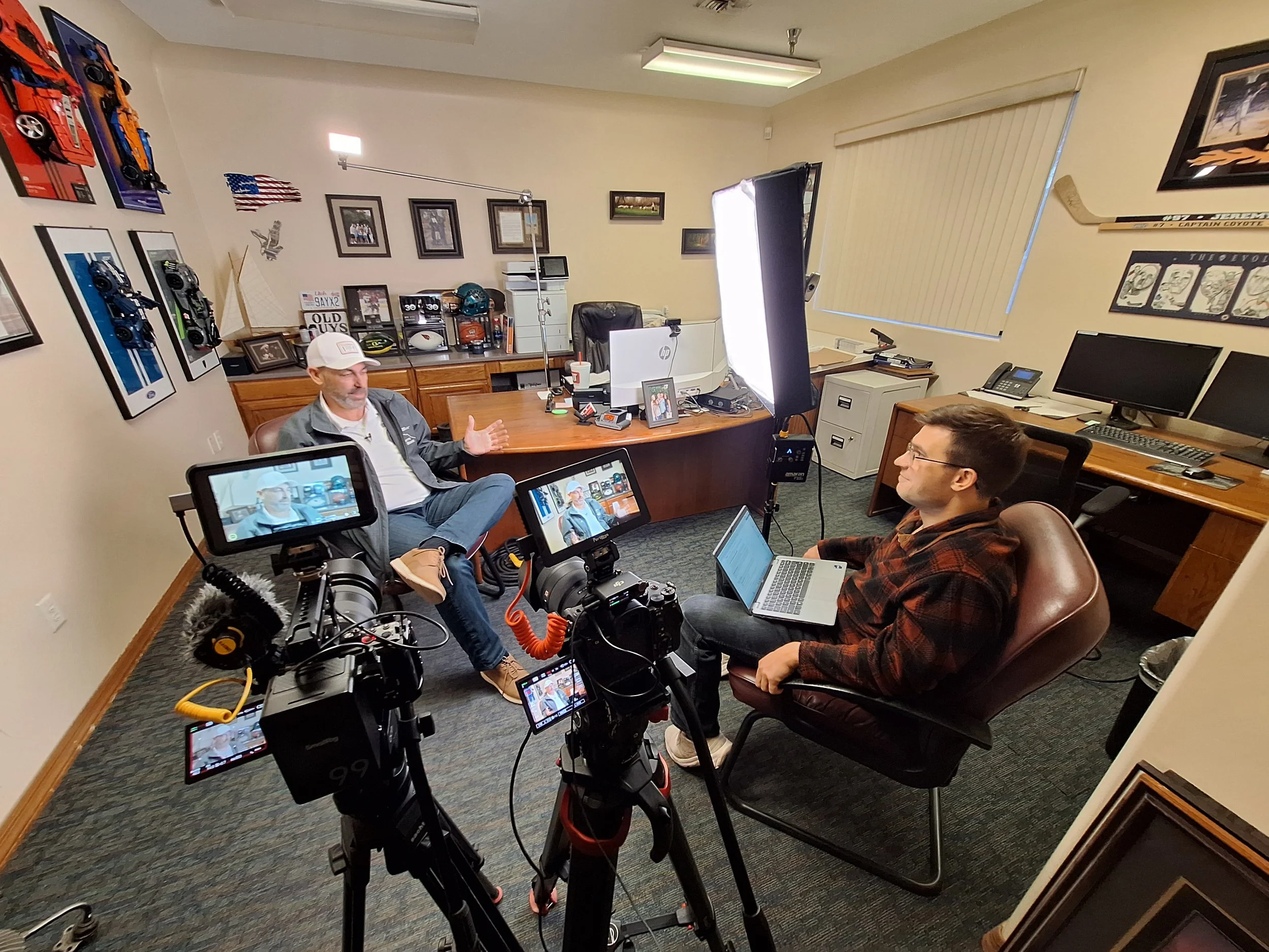 Two men are seated in an office, one speaking and the other listening with a laptop. Two cameras are recording the interview, with professional lighting and office decor visible.