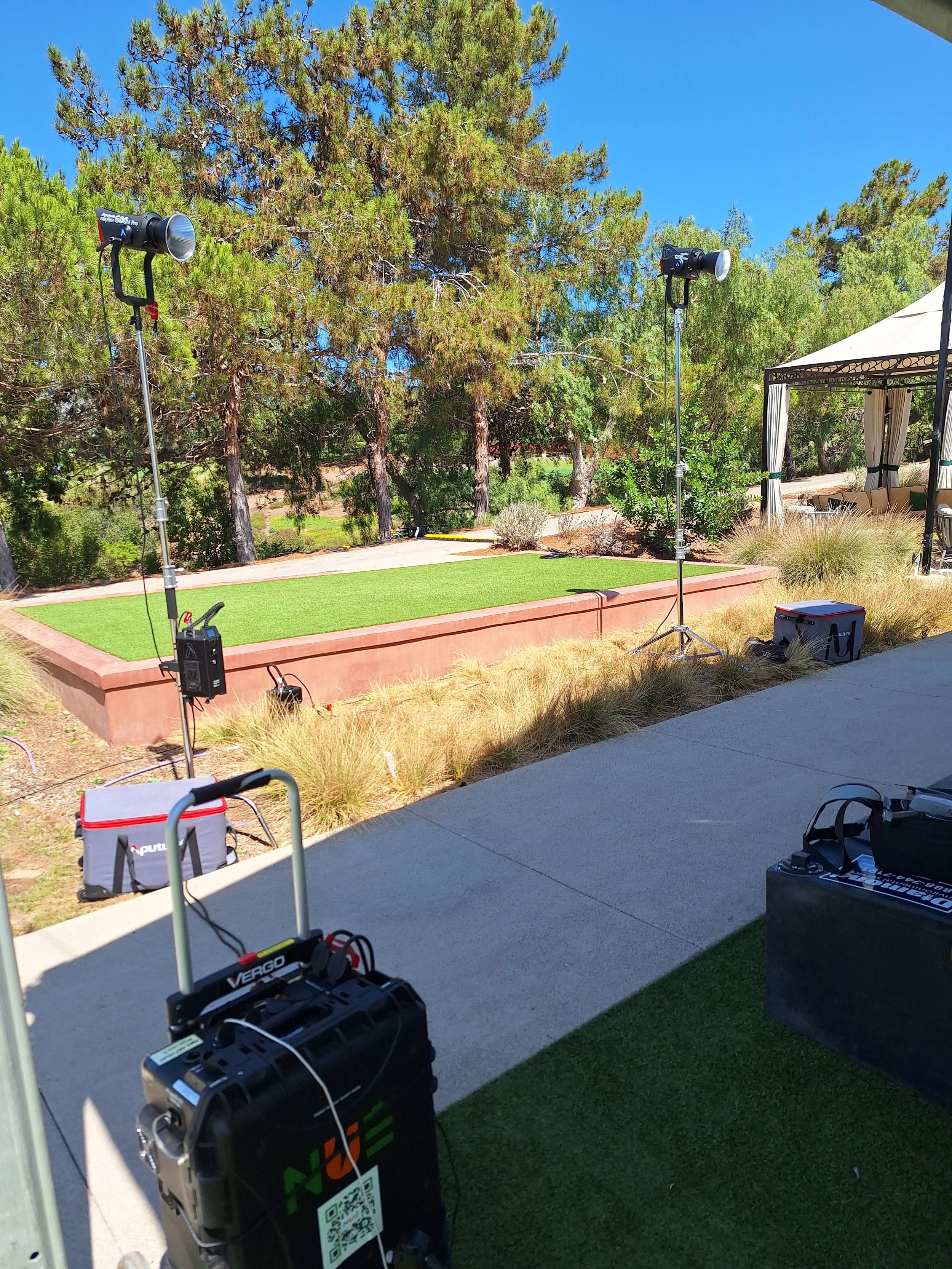 Stage setup with two large professional lights on tall stands directed towards a small artificial grass stage in an outdoor area with trees and a tent, indicating a setup for a photoshoot or filming outdoors.