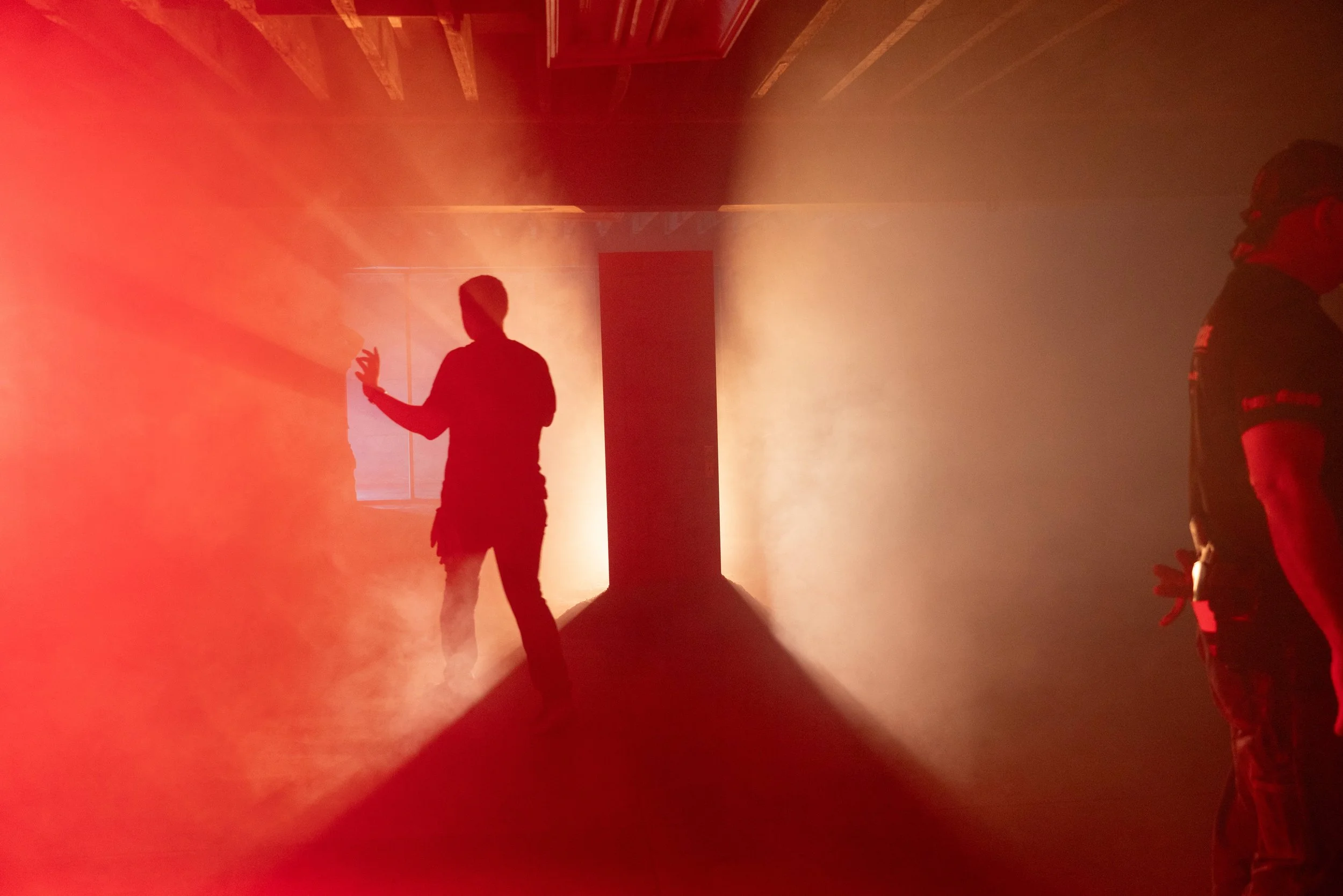 Silhouettes of two people standing in a hallway with fog and red-orange lighting, one person gesturing with their hand, the other partially visible.
