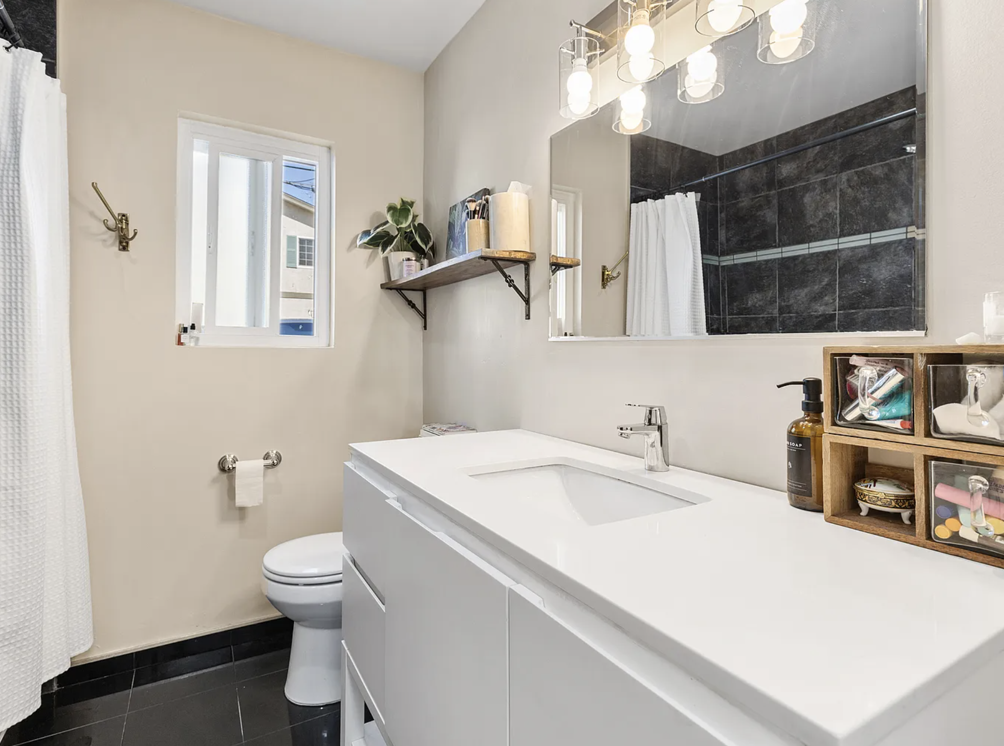 Modern bathroom with white vanity, large mirror, toilet, wall-mounted shelf, and decorative lighting.
