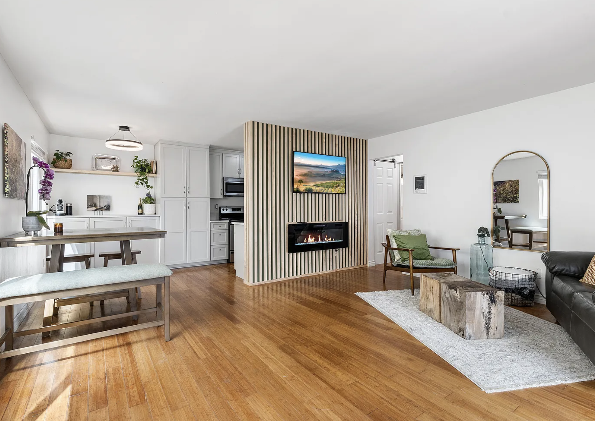 Modern living room and kitchen with wooden floors, white cabinets, a dining table, and a cozy sitting area with a fireplace.