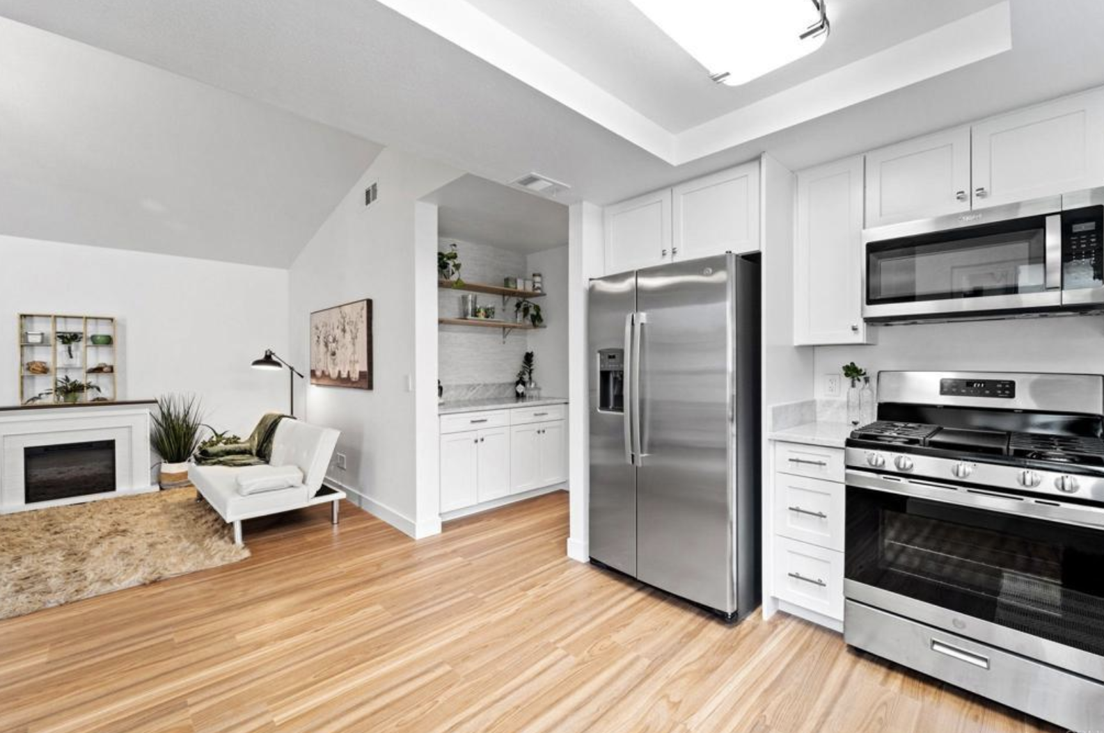 Modern kitchen with stainless steel appliances including a refrigerator and oven, white cabinetry, and a small adjacent living area with a chair and potted plants.